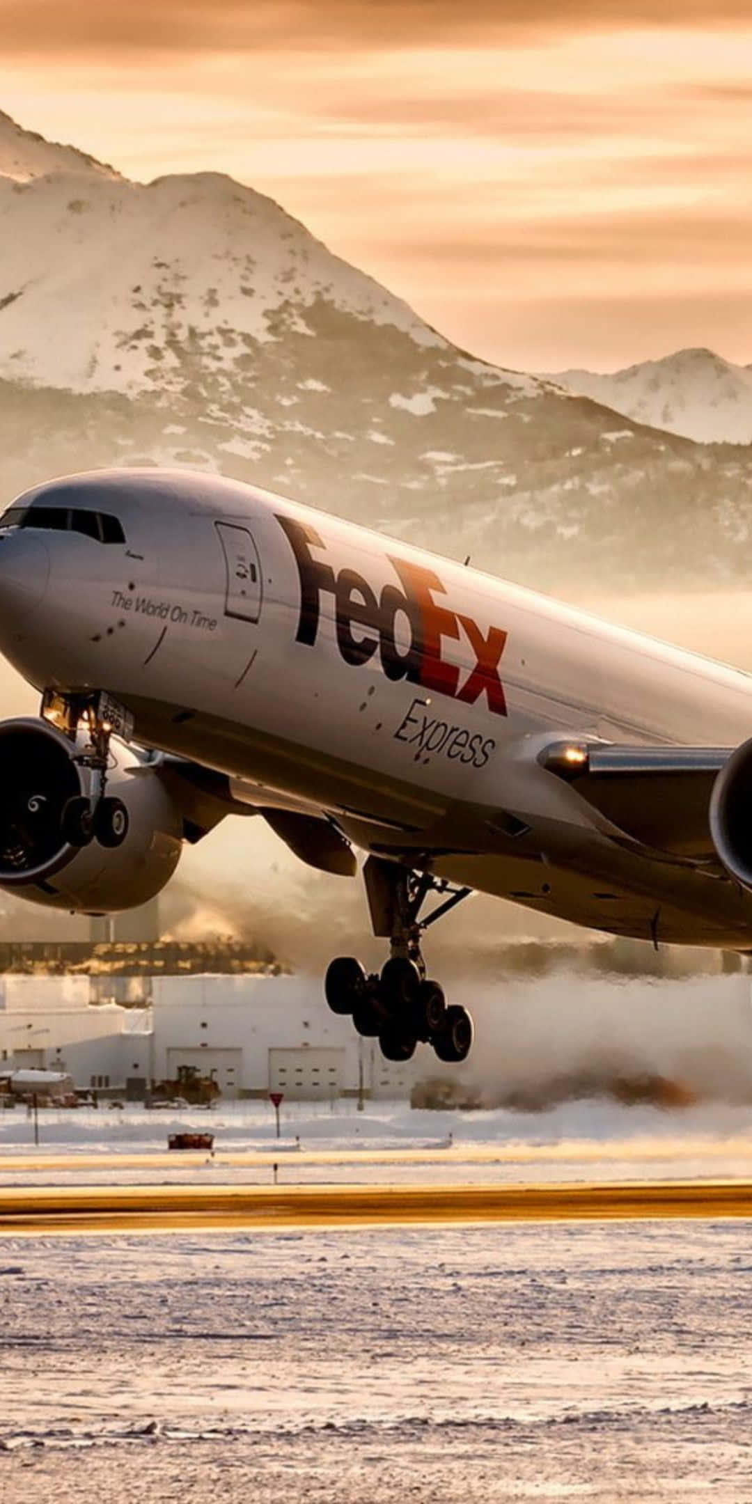 天空之美 壁紙 — 一架 FedEx Express 貨機從積雪跑道起飛，白雪覆蓋的山峰在金色夕陽下閃耀。