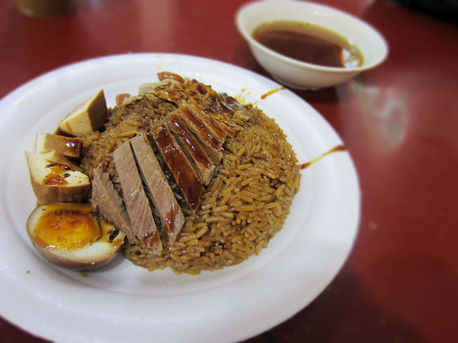 天籟新加坡鴨飯斜角拍攝 壁紙 — 一碟油亮烤鴨配糙米飯、豆腐和半邊蛋，旁邊有一小碗醬汁，放在紅色桌子上。