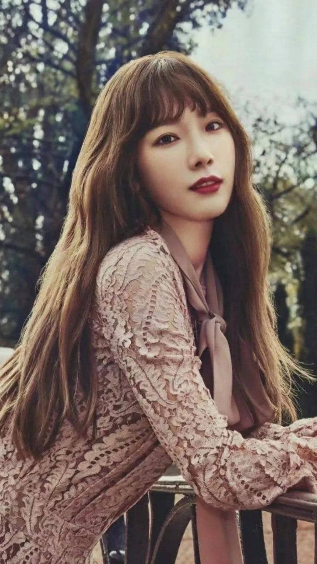太妍林中風情 壁紙 — 一位擁有長長棕色頭髮和紅唇的女性，在樹林環繞的戶外環境中倚靠在欄杆上，身穿粉紅色蕾絲上衣。