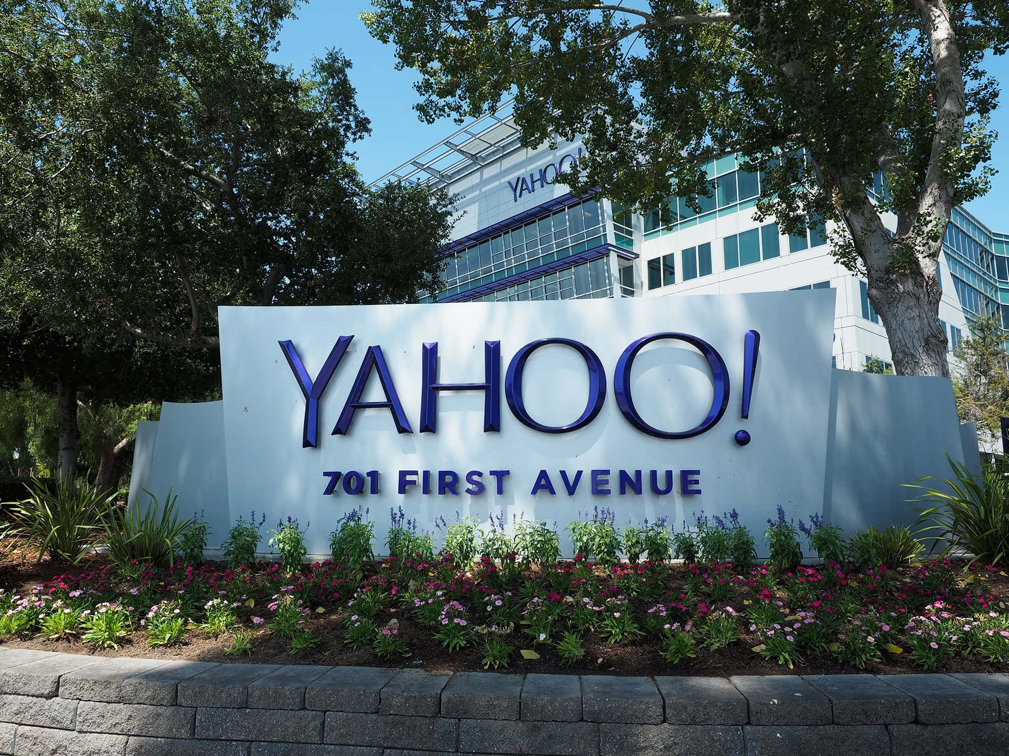 太平洋上空壯麗日落 壁紙 — 位於701 First Avenue的大型藍色Yahoo標誌，被綠樹和色彩繽紛的花朵環繞，在晴朗的藍天下。