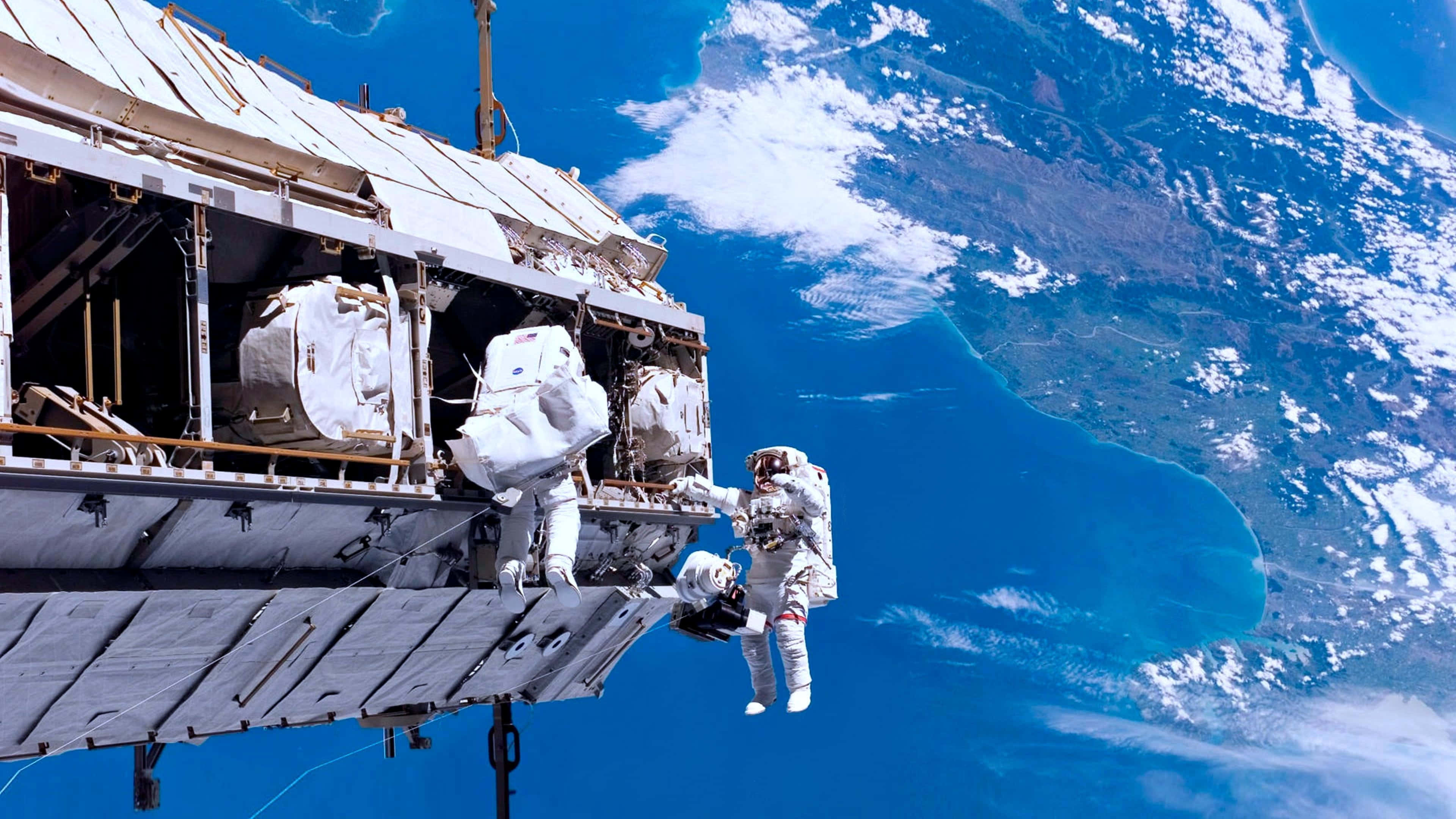 Space Technology 桌布 壁紙 — 太空人在太空站外進行太空漫步，下方可見地球的藍色海洋和白色雲層。