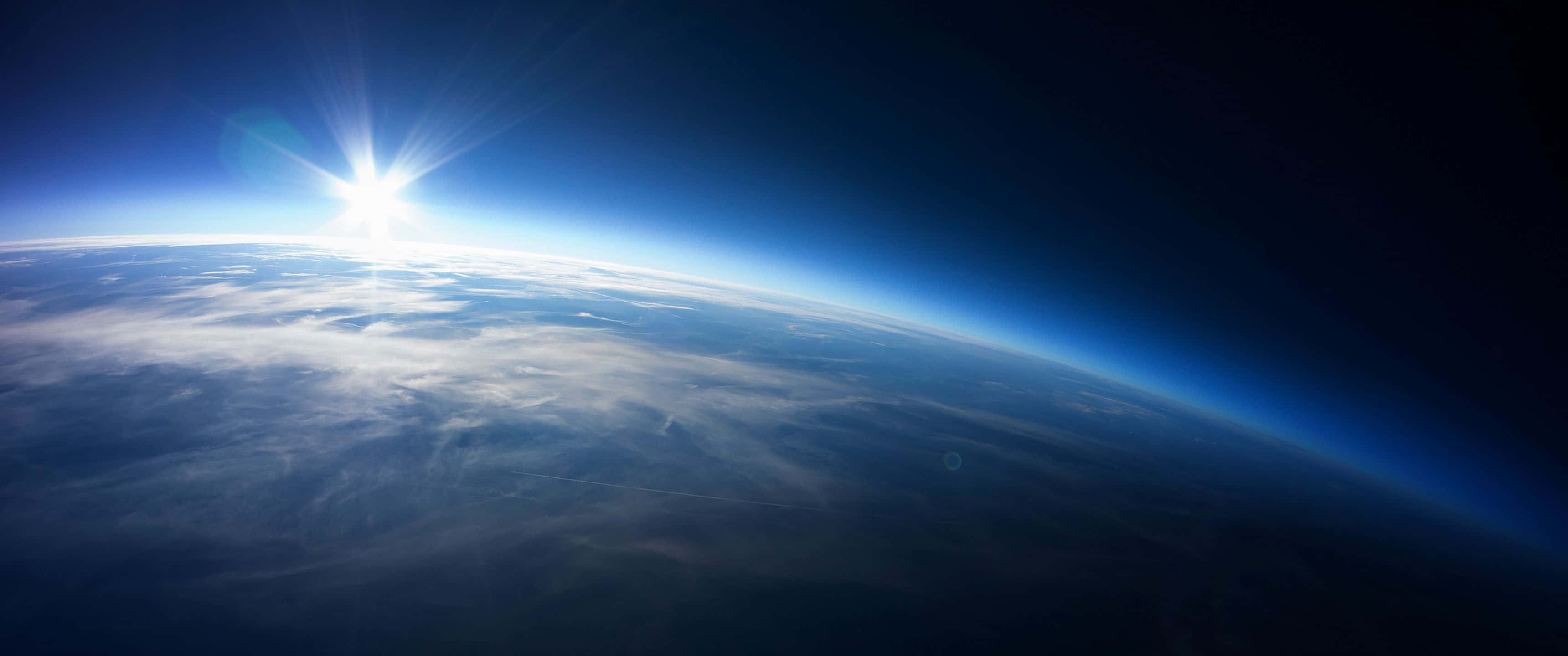 太空日出 3440x1440p 背景 壁紙 — 從太空看地球上的日出，發光的地平線和下方的雲層。