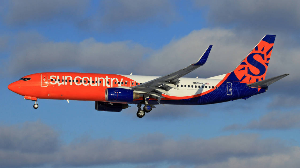 Sun Country Airlines 的噴射機以橙色和藍色塗裝，飛行在部分多雲的天空中。