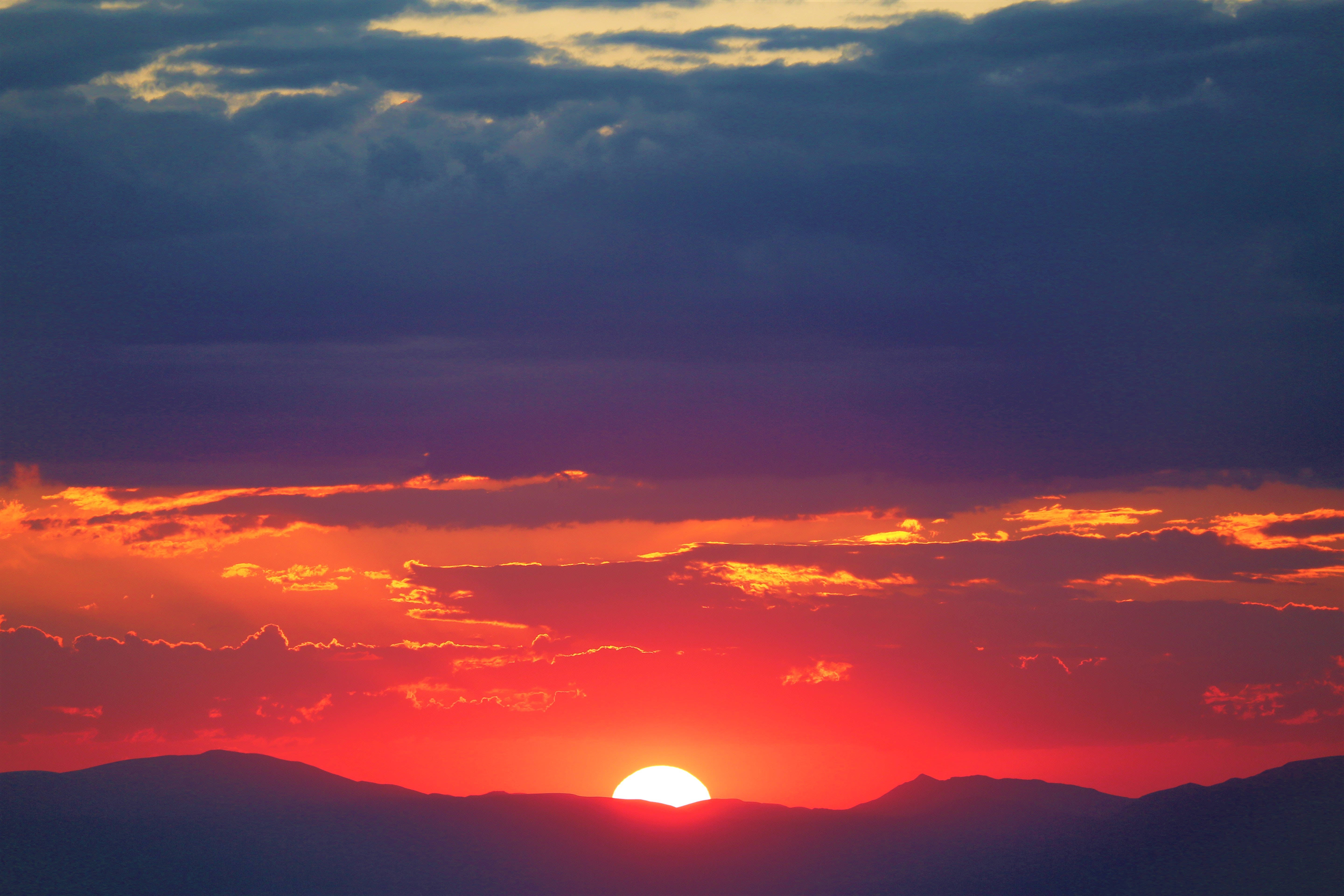 太陽從山巒間透出的唯美天空 壁紙 — 熾熱的夕陽將天空染成鮮豔的紅色和紫色，太陽在輪廓山脈後方落下。
