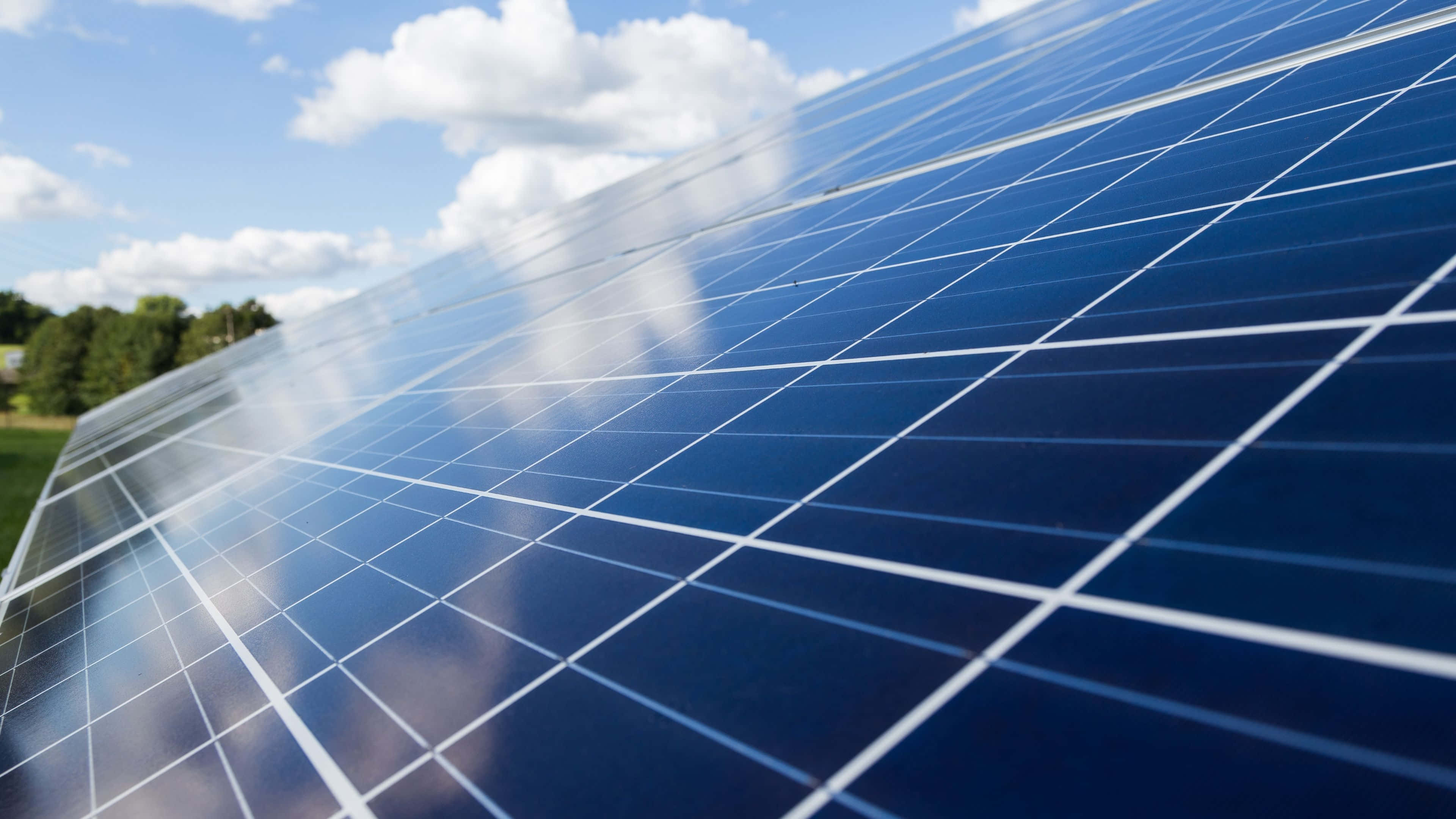Solar Panel 桌布 壁紙 — 太陽能板特寫，反射出明亮的藍天、蓬鬆的白雲和背景中的綠樹。