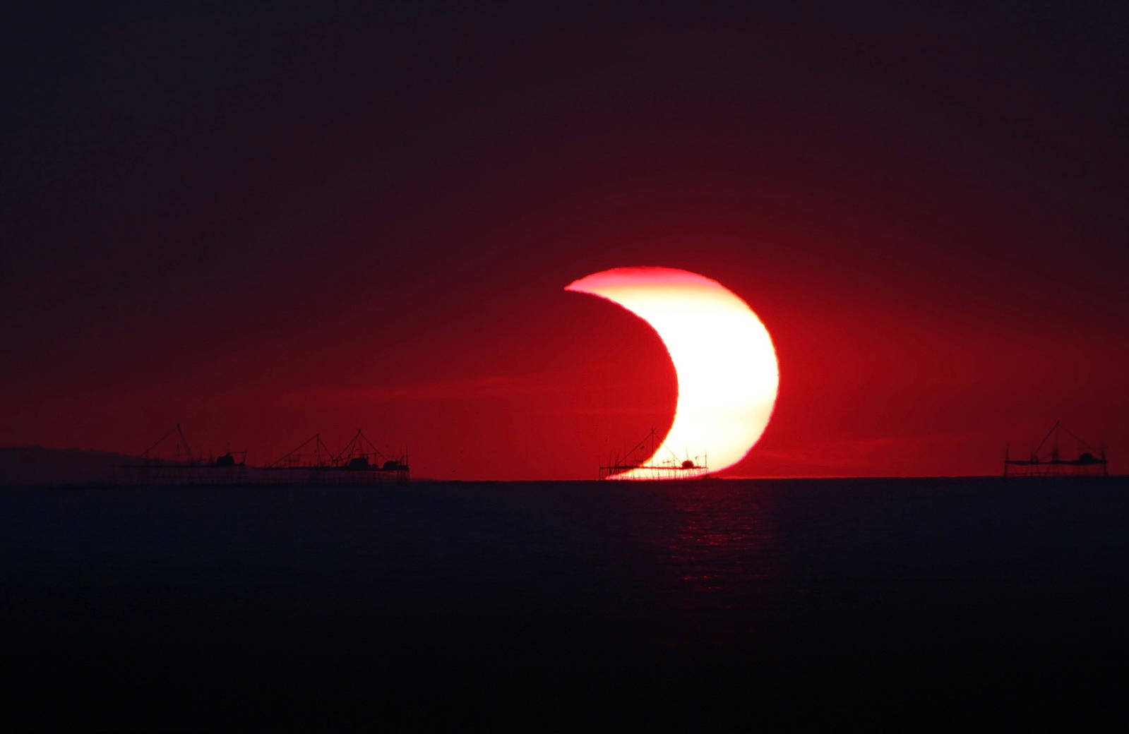 太陽與月亮擁抱於紅天 壁紙 — 戲劇性的紅色新月太陽在黑暗的海洋上發生食，漁具的剪影映襯在深紅色的天空中。
