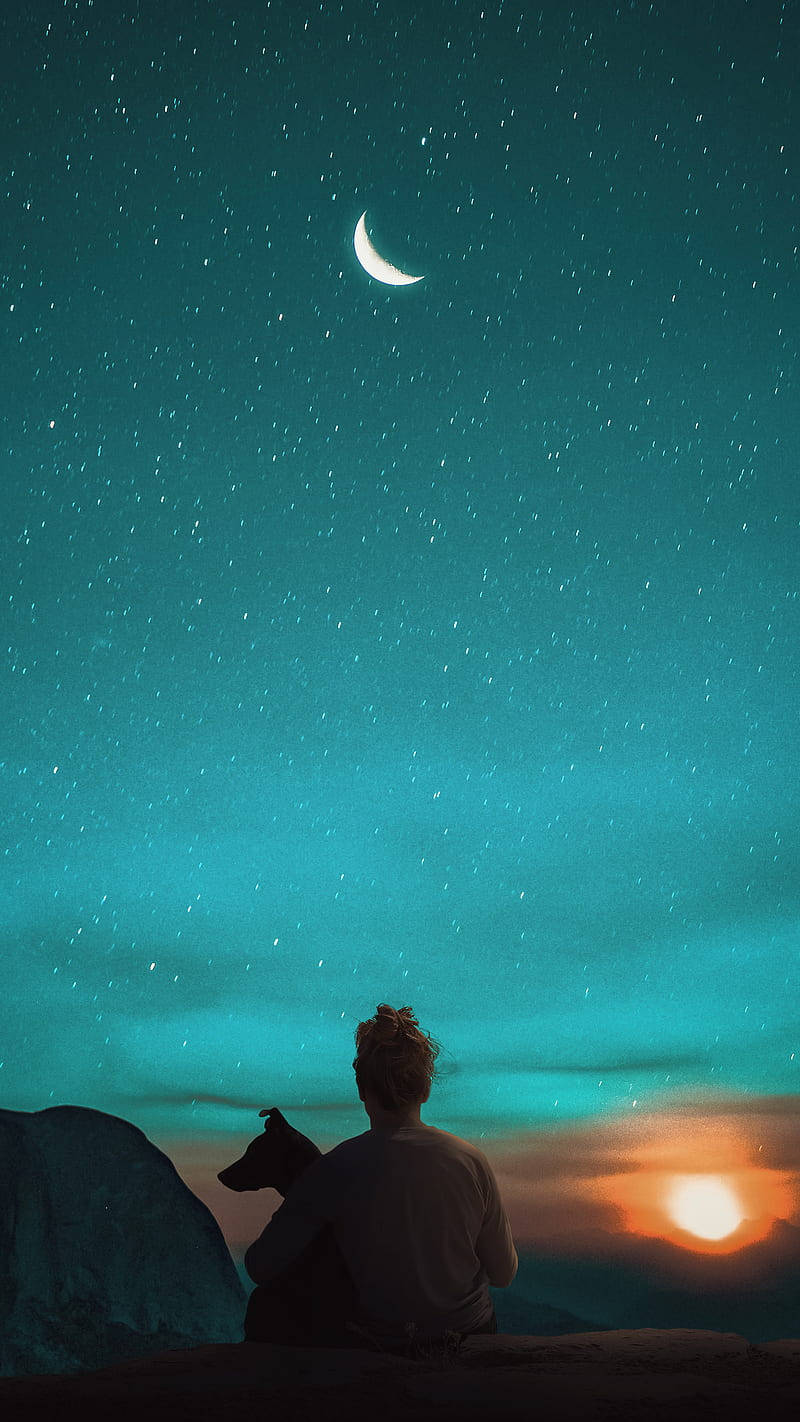 太陽與月亮於碧藍天空 壁紙 — 一個人同一隻狗以剪影形式坐喺懸崖上，觀賞住一彎新月喺佈滿星星嘅藍綠色天空升起，而太陽喺下面日落。