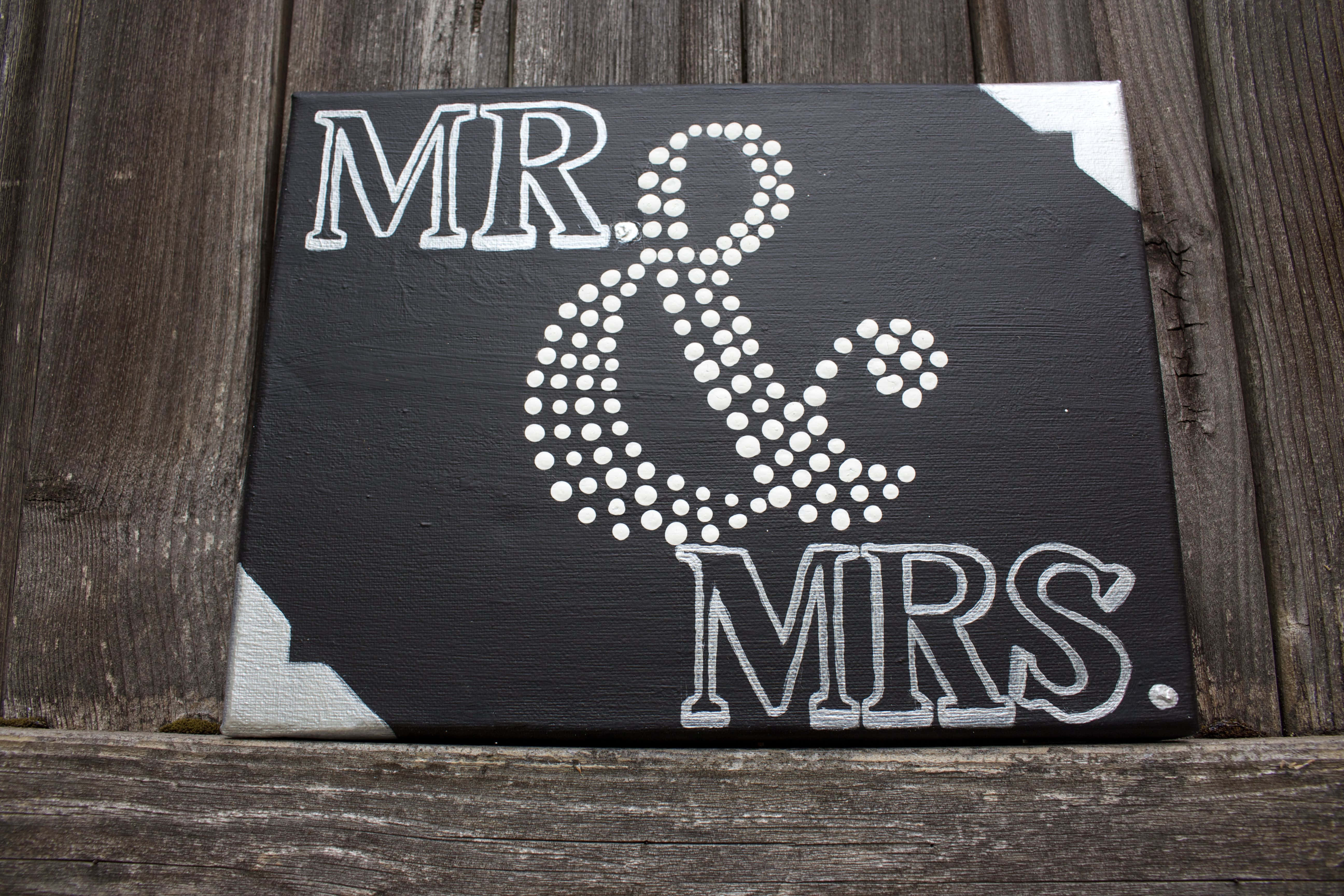 黑色畫布上印有白色 'MR & MRS' 文字及圓點和號，木質表面。