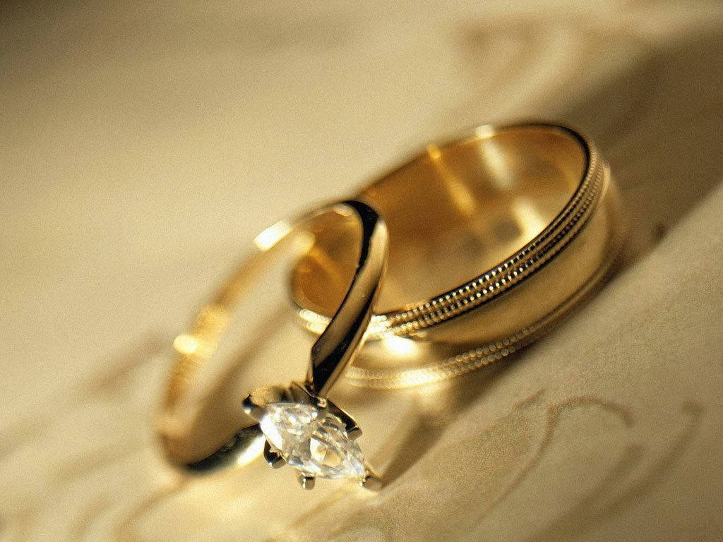 夫婦婚戒 壁紙 — 兩枚金結婚戒指，其中一枚鑲有馬眼形鑽石，放在柔軟的奶油色布料上，沐浴在溫暖的金色光線中。