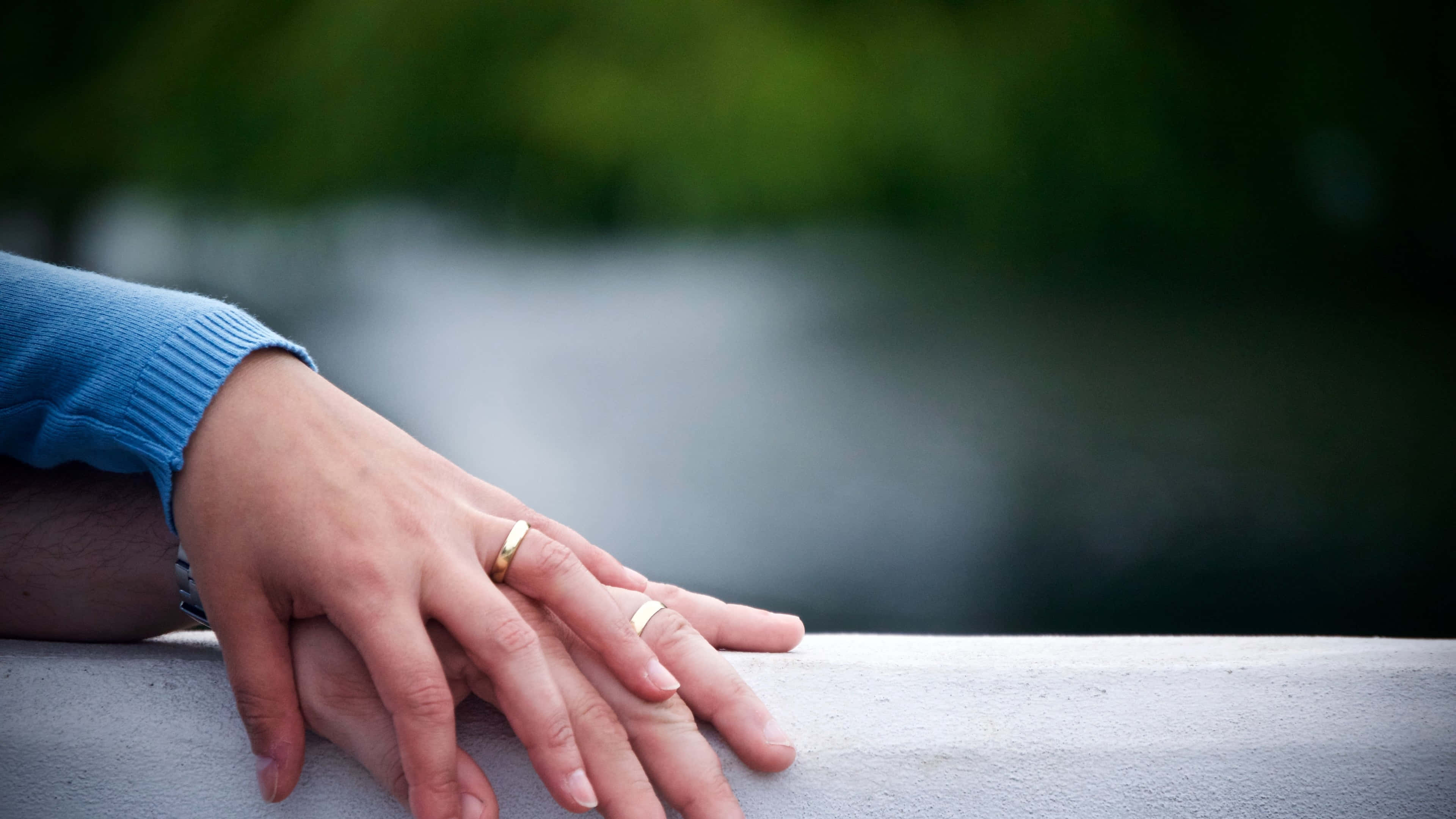 夫婦婚戒握於欄杆 壁紙 — 兩隻手緊握在一起，戴著結婚戒指，擱在石頭邊緣上，背景是模糊的綠色和灰色。