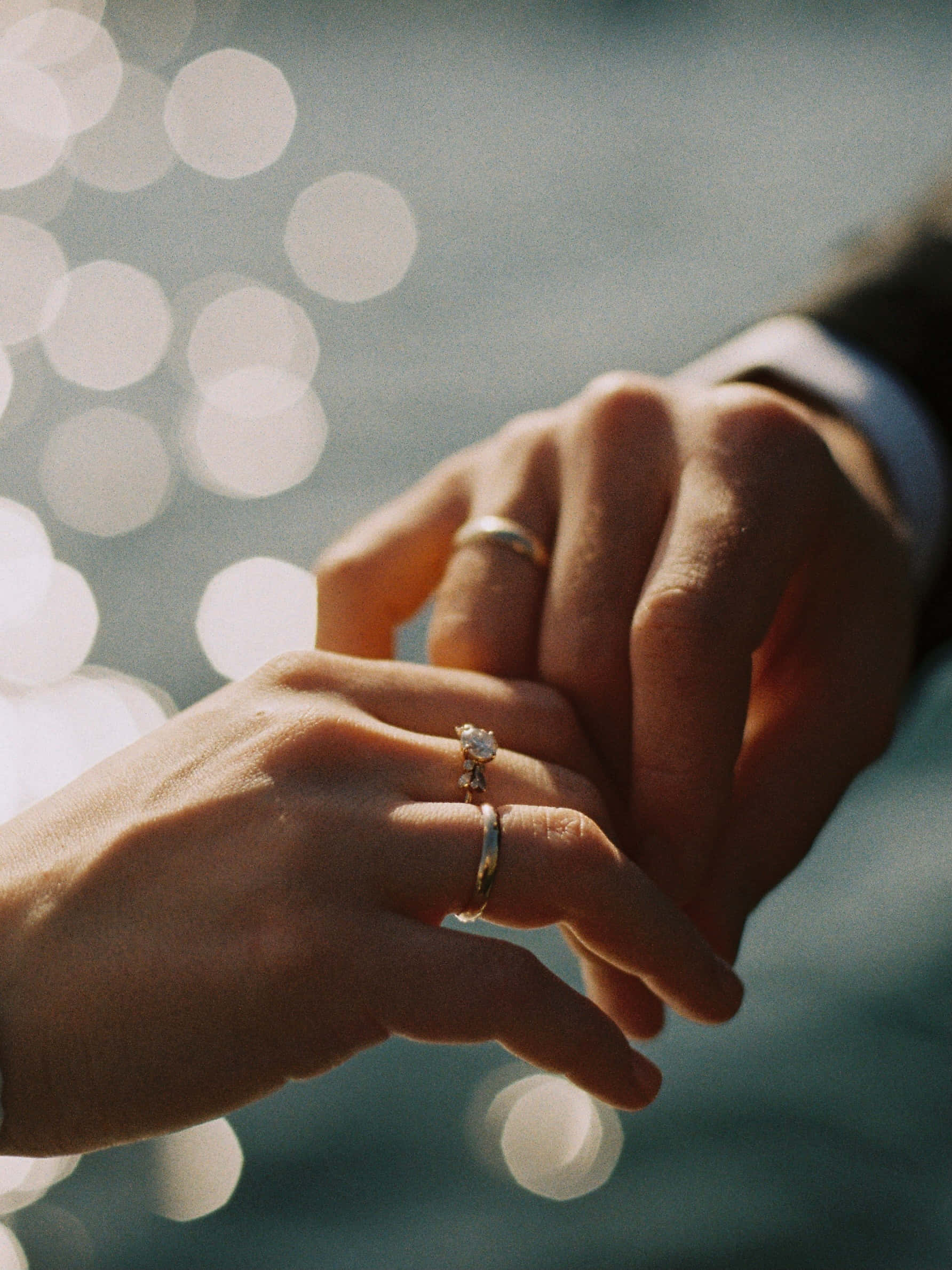 夫婦擁抱展示婚戒 壁紙 — 兩隻手輕輕交握，戴著結婚戒指，背景是柔和的水面陽光散景。