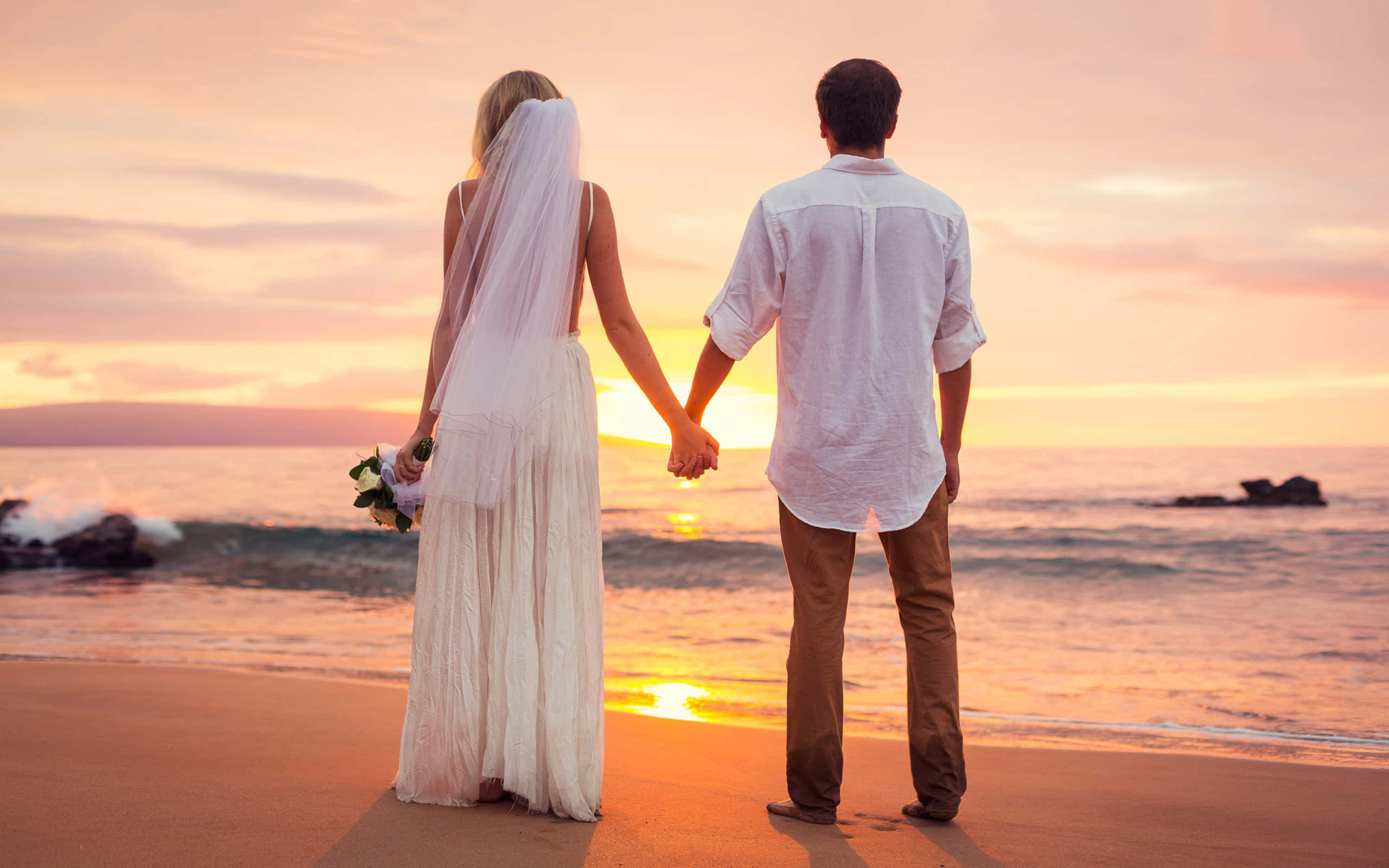 夫婦海灘日落婚禮 壁紙 — 新娘和新郎在海灘上牽手，夕陽以溫暖的橙色和粉紅色調在海洋上空落下。