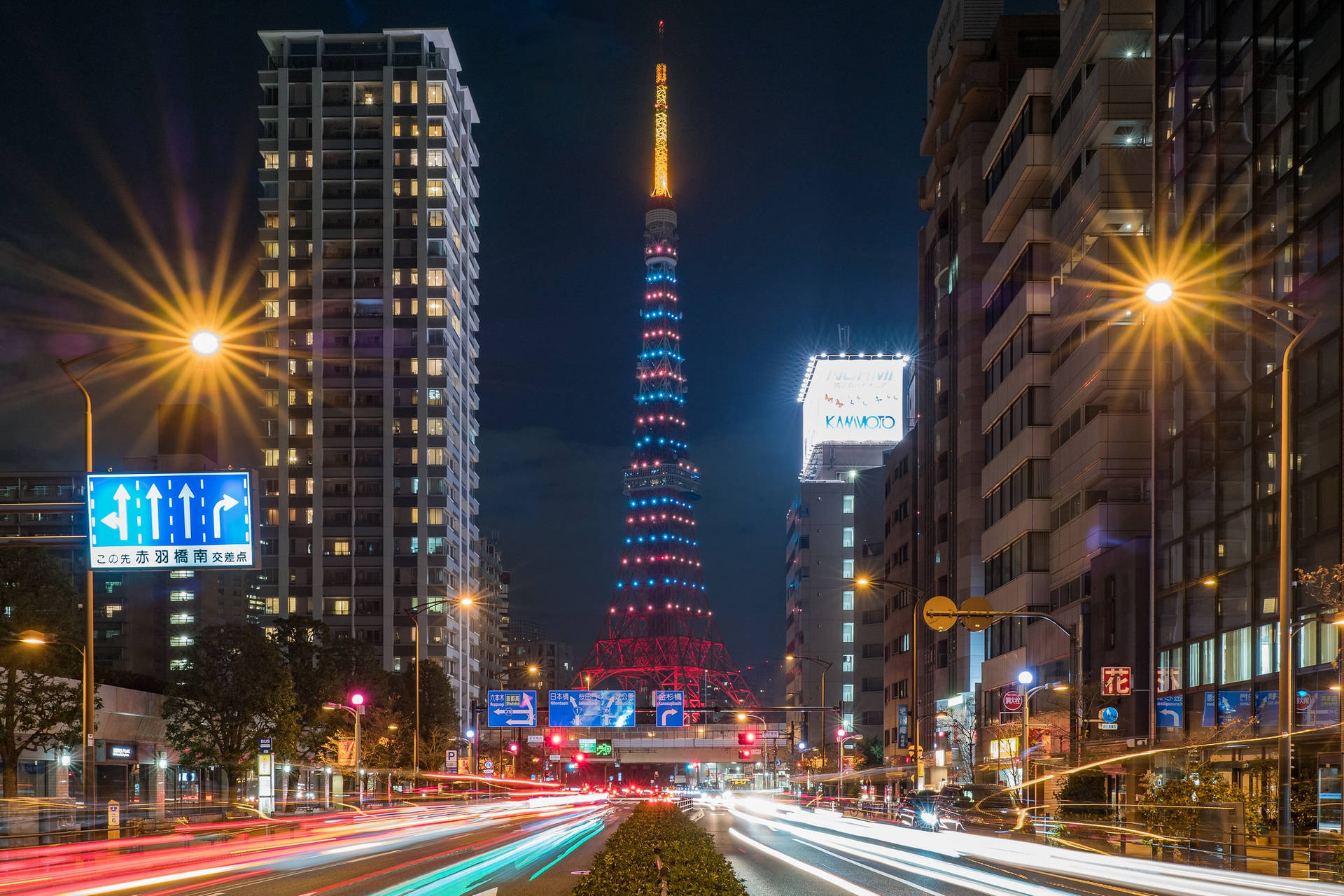 夾在建築間的東京塔 壁紙 — 充滿活力的夜景，東京塔閃爍著紅、藍、黃光，被高樓大廈和汽車燈光軌跡環繞。