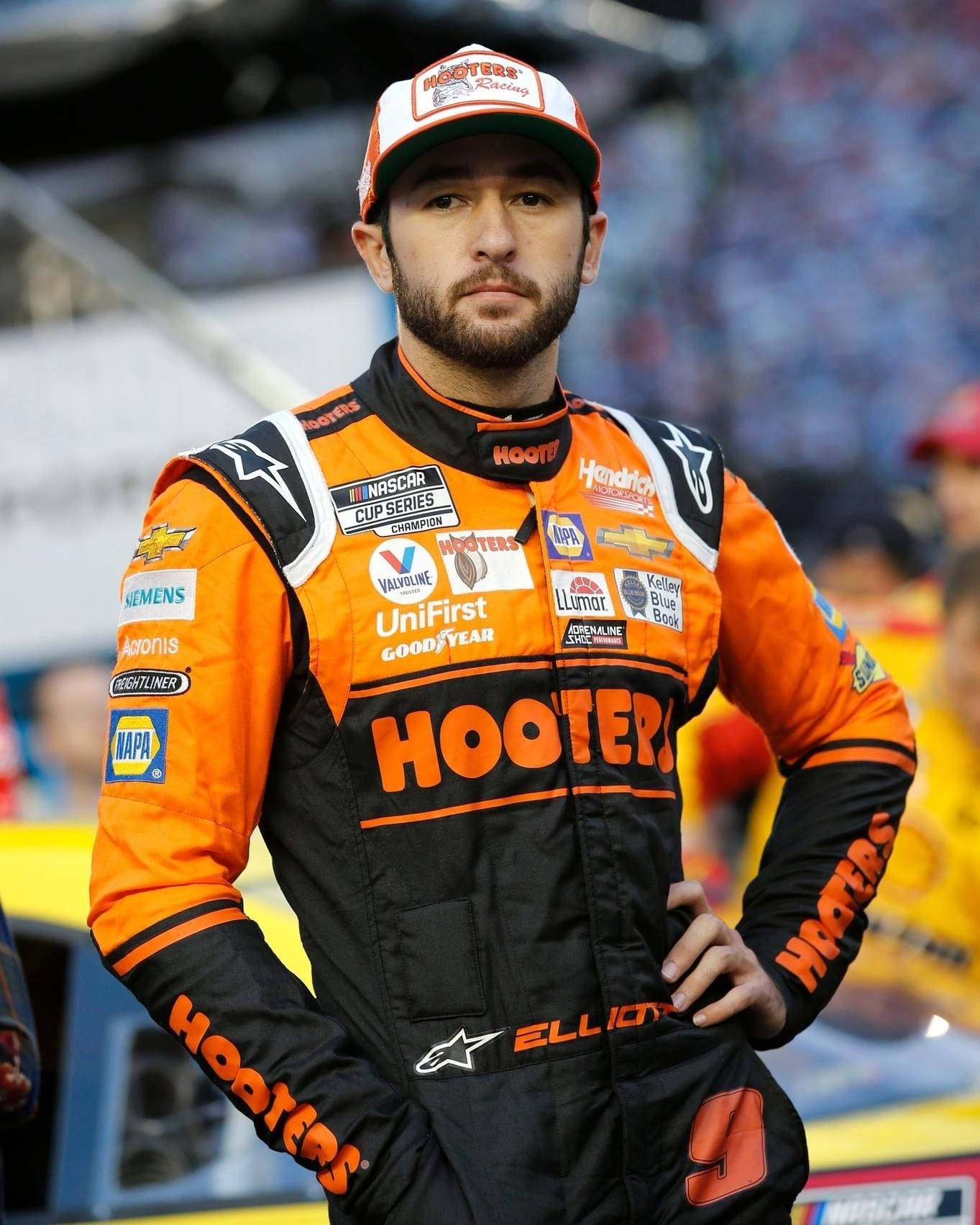 身穿橙色和黑色Hooters賽車服的NASCAR車手在賽道上自信地站立。
