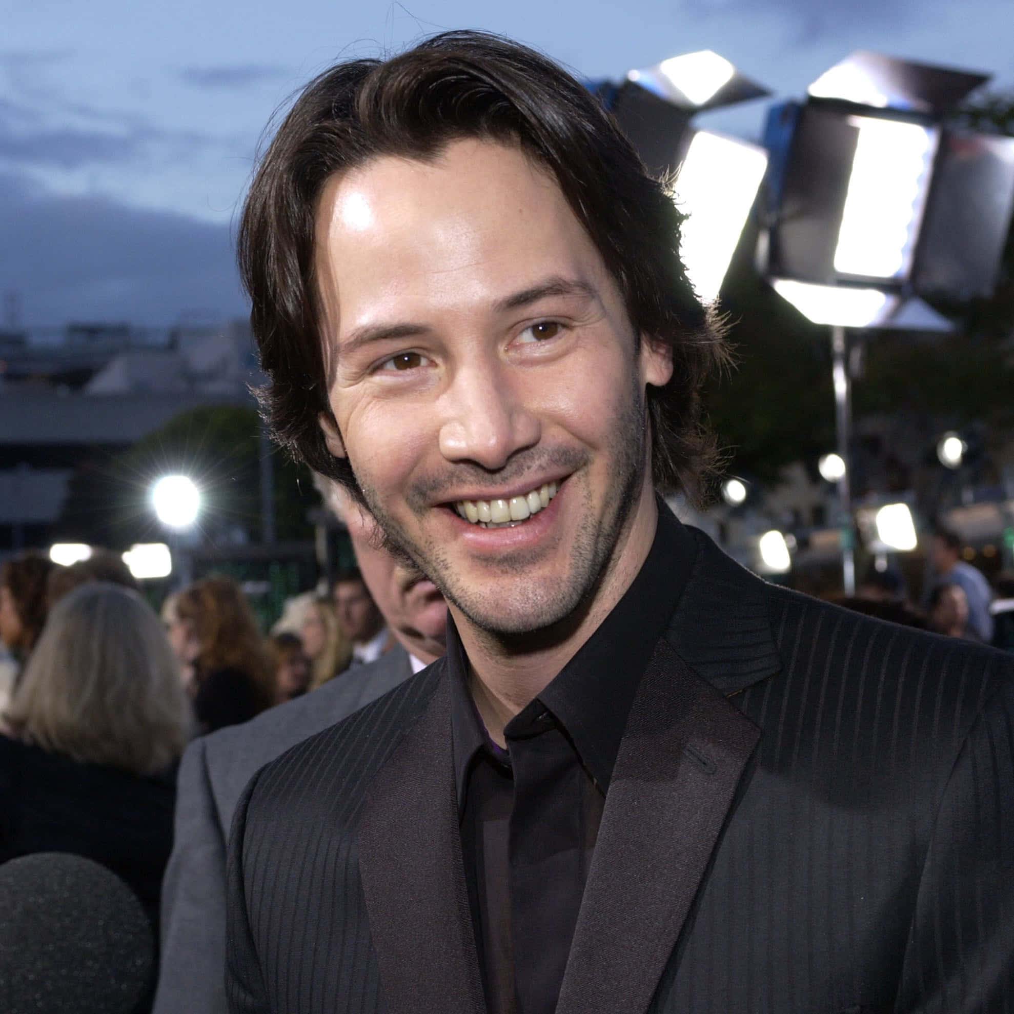 Keanu Reeves 桌布 壁紙 — 奇洛·李維斯在黃昏的明亮活動燈光下穿著細條紋西裝溫暖地微笑。