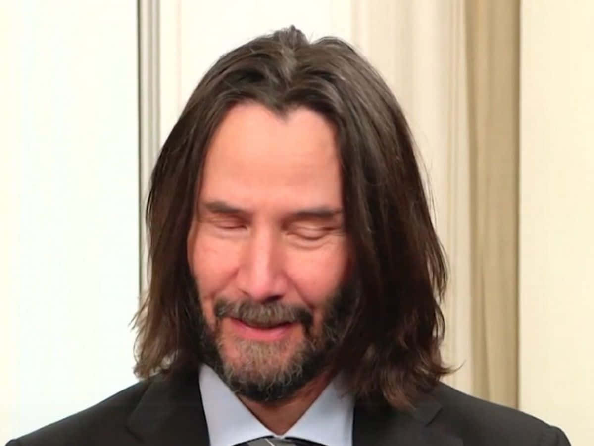 Keanu Reeves 桌布 壁紙 — 奇洛·李維斯留著長黑髮和鬍鬚，在淺色背景前穿著黑色西裝打領帶，溫暖地微笑。