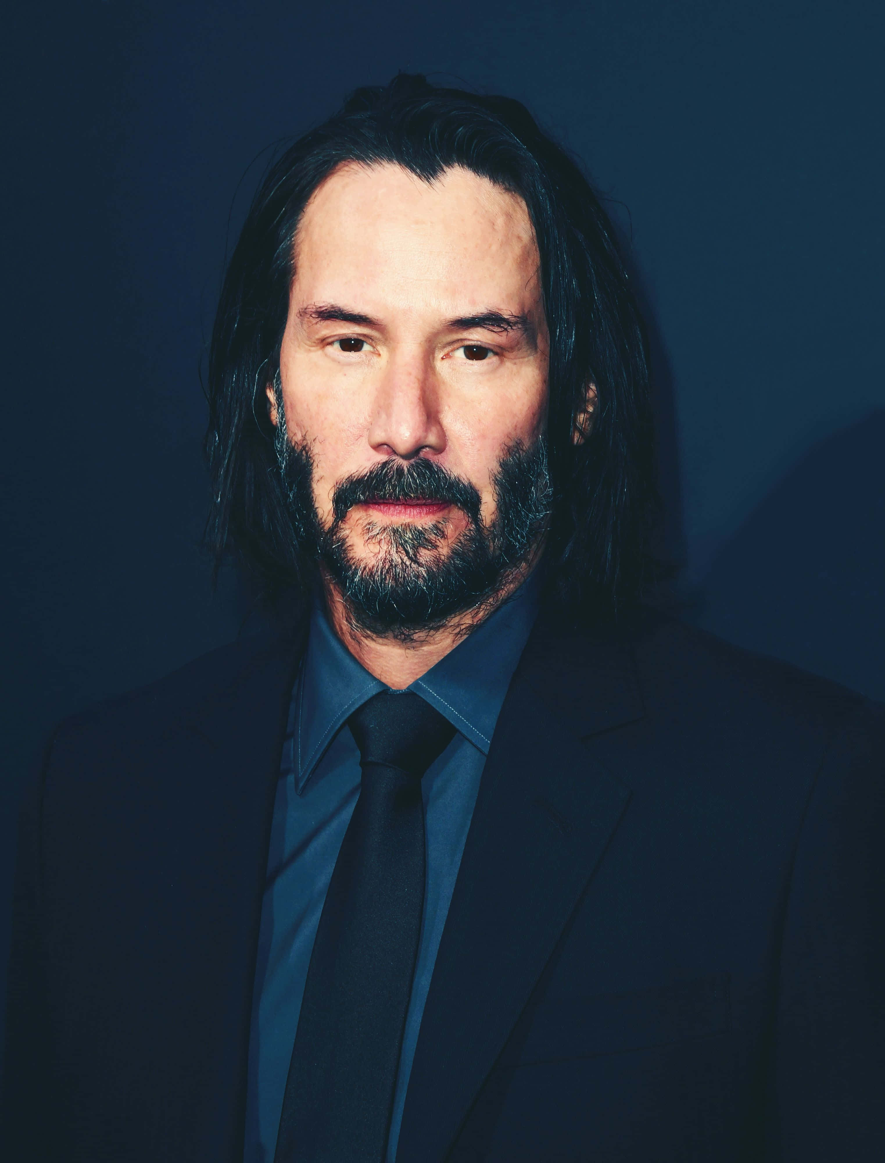 Keanu Reeves 桌布 壁紙 — 奇洛·李維斯的嚴肅肖像，長黑髮和鬍鬚，身穿深色西裝，背景為深藍色。