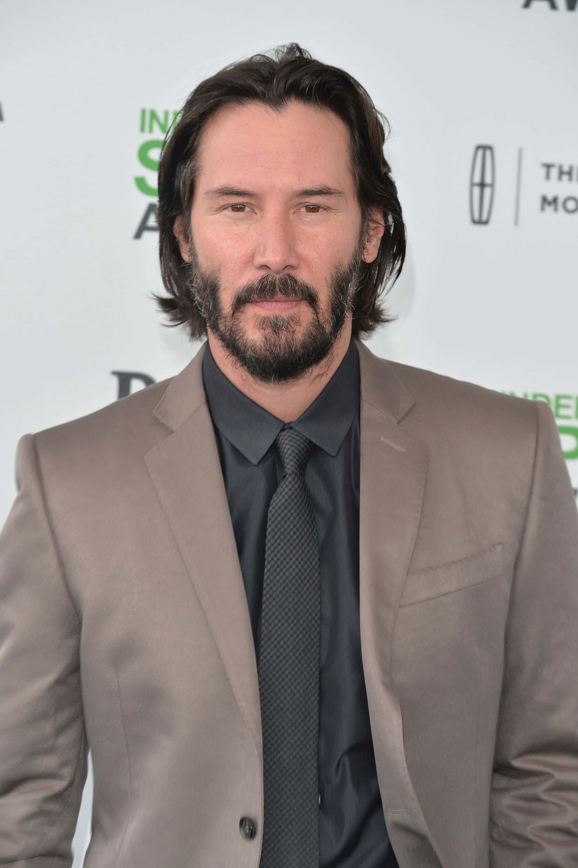 Keanu Reeves 桌布 壁紙 — 奇洛·李維斯穿著灰褐色西裝和深色領帶在紅地毯活動上擺姿勢，身後有贊助商標誌。