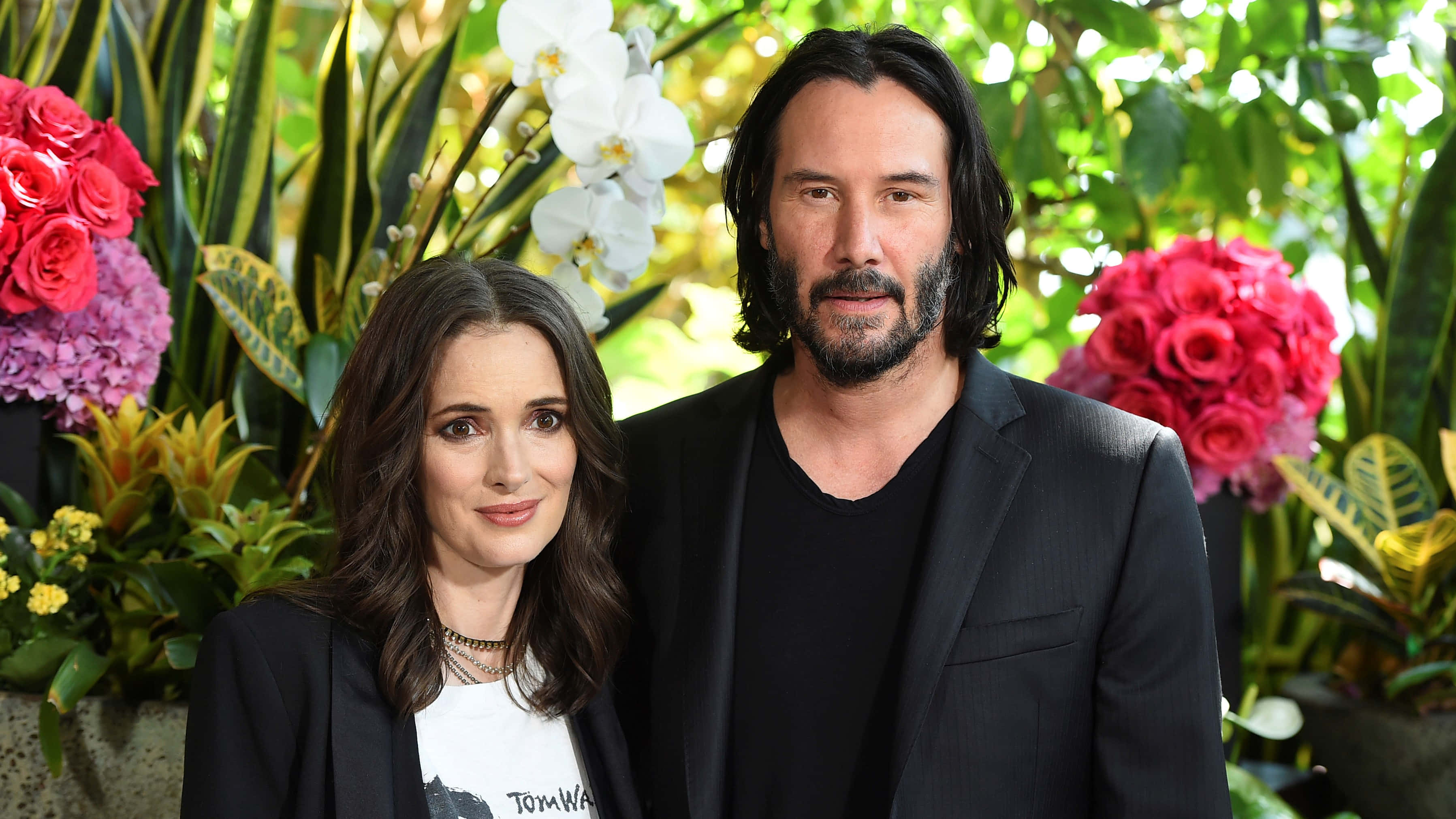 Keanu Reeves 桌布 壁紙 — 奇洛·李維斯與雲露娜·維達在明媚日光下，置身於茂盛的熱帶花卉與綠植中一同擺姿勢。