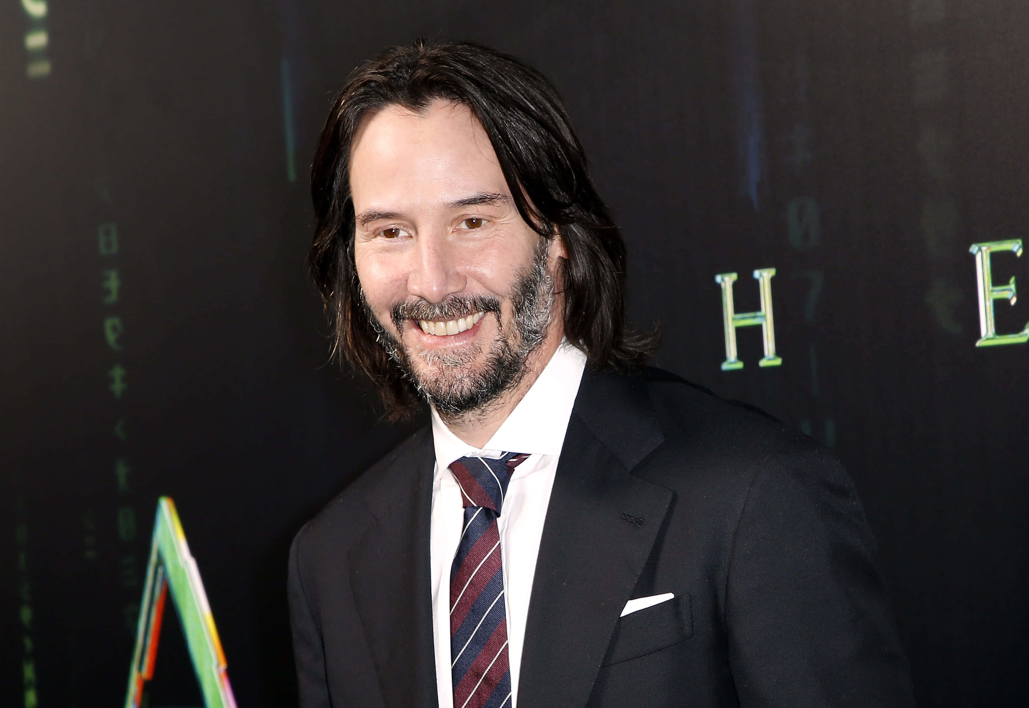 Keanu Reeves 桌布 壁紙 — 奇洛·李維斯身穿利落黑色西裝，在黑暗背景中散發著綠色發光矩陣代碼和字母，溫暖地微笑。