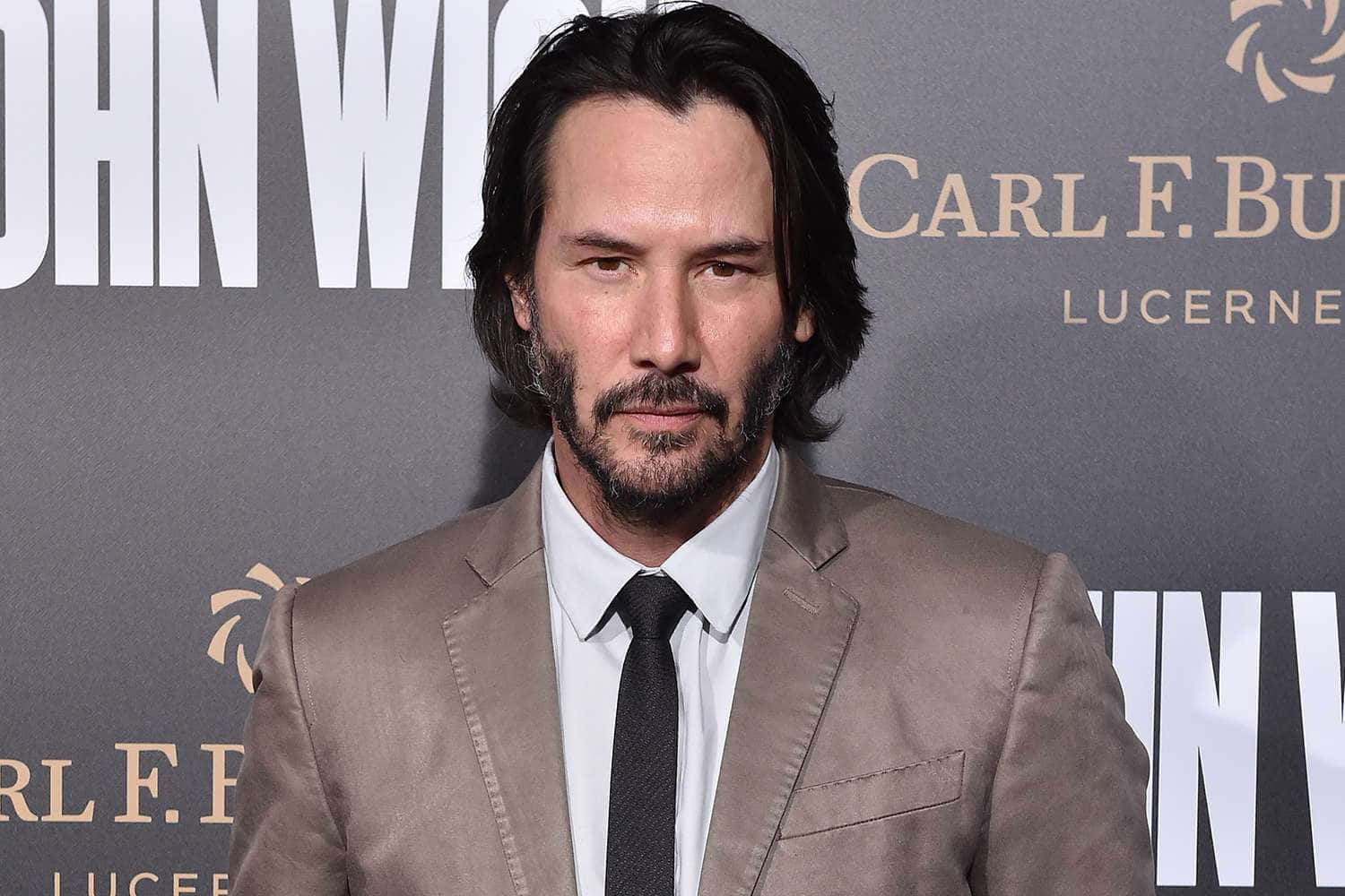 Keanu Reeves 桌布 壁紙 — 奇洛·李維斯身穿米色西裝和黑色領帶，在帶有品牌背景的《殺神John Wick》首映活動上嚴肅擺姿勢。