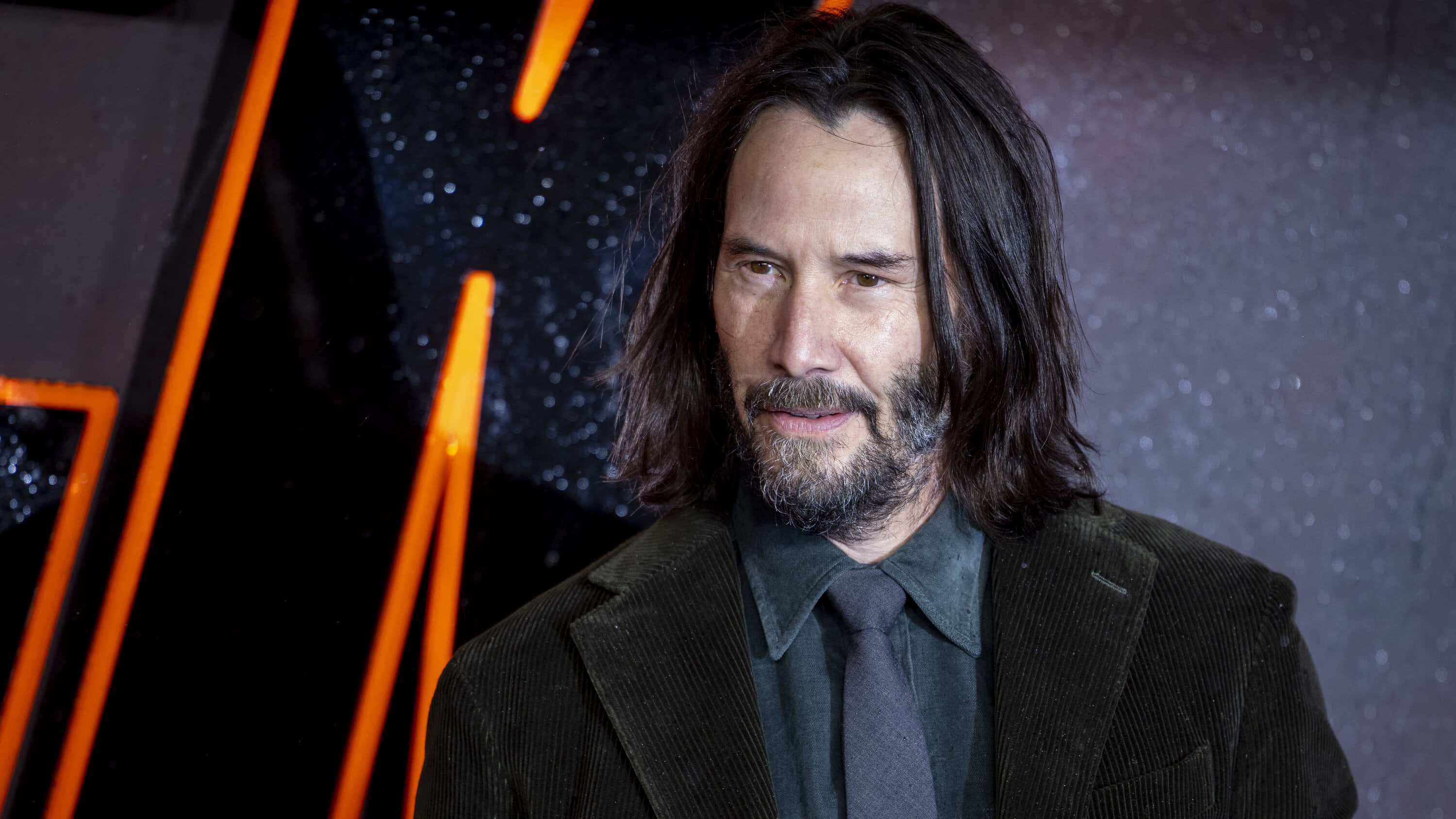 Keanu Reeves 桌布 壁紙 — 奇洛李維斯留著長黑髮和鬍鬚，身穿深色燈芯絨西裝，背景是星空和發光的橙色線條。