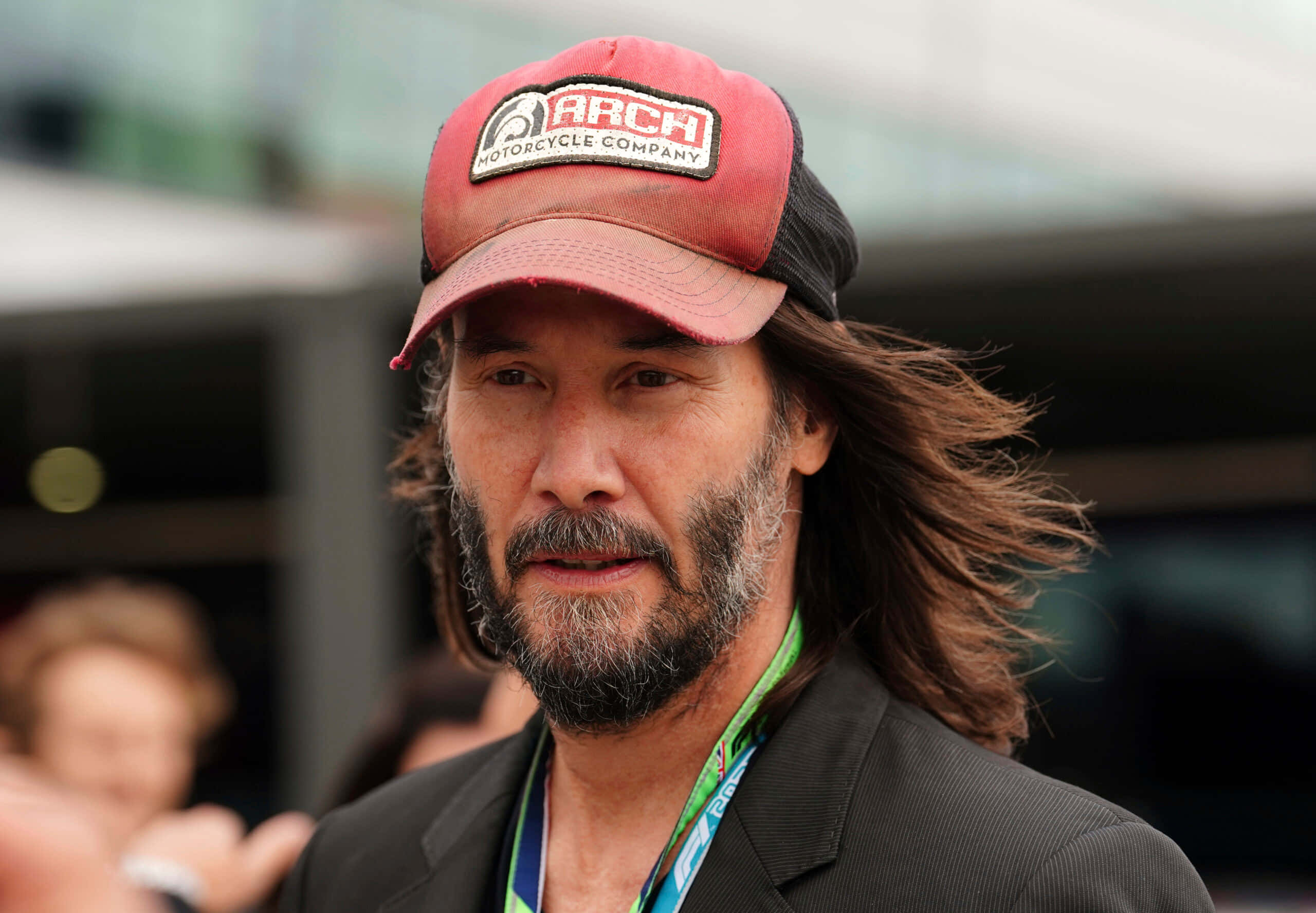 Keanu Reeves 桌布 壁紙 — 奇洛李維斯留長髮和鬍鬚，在戶外戴著紅色ARCH Motorcycle Company帽子和深色西裝。