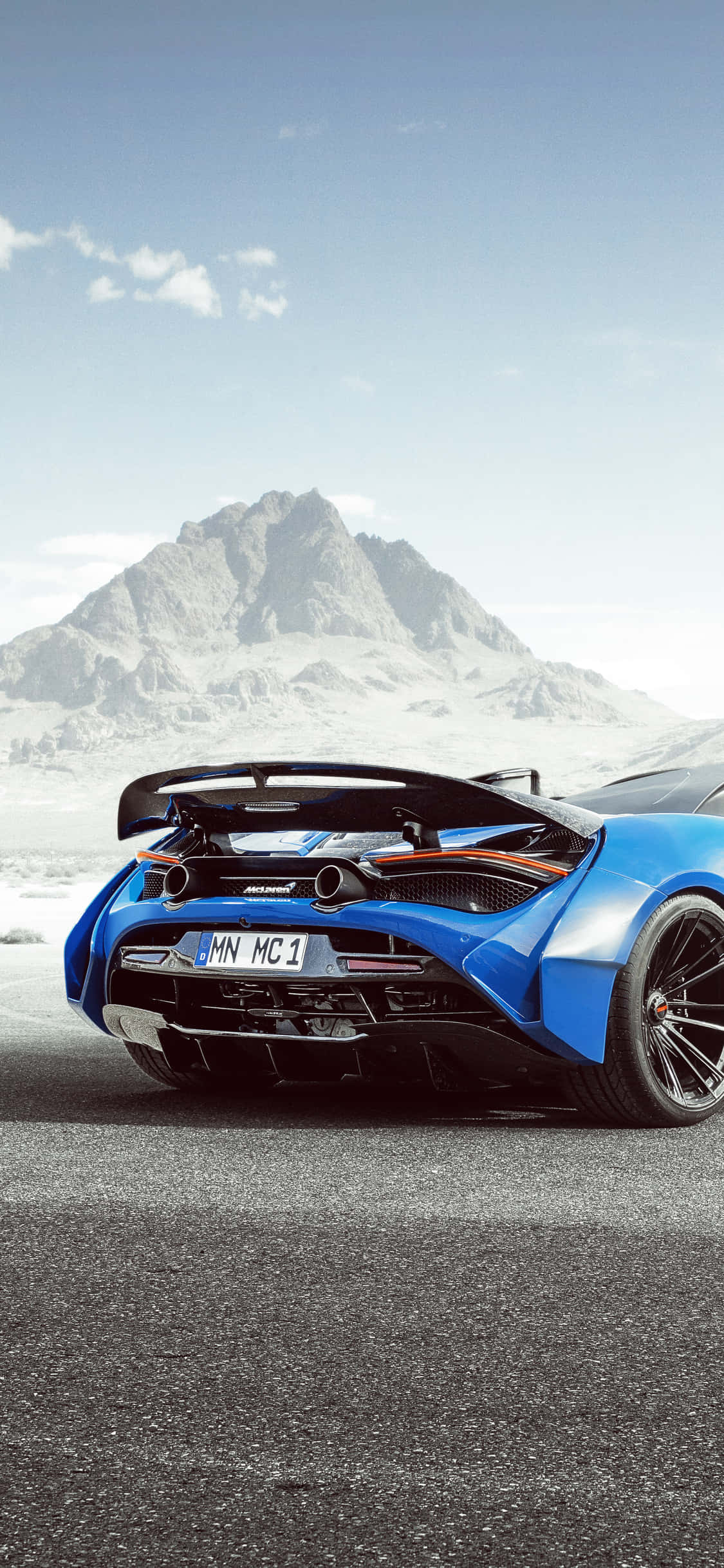 奢華 iPhone Xs 與 McLaren 720S 同框呈現 壁紙 — 一輛鮮藍色McLaren跑車，帶有大型後擾流板，停在瀝青路面上，背景是白雪覆蓋的山脈，天空晴朗。