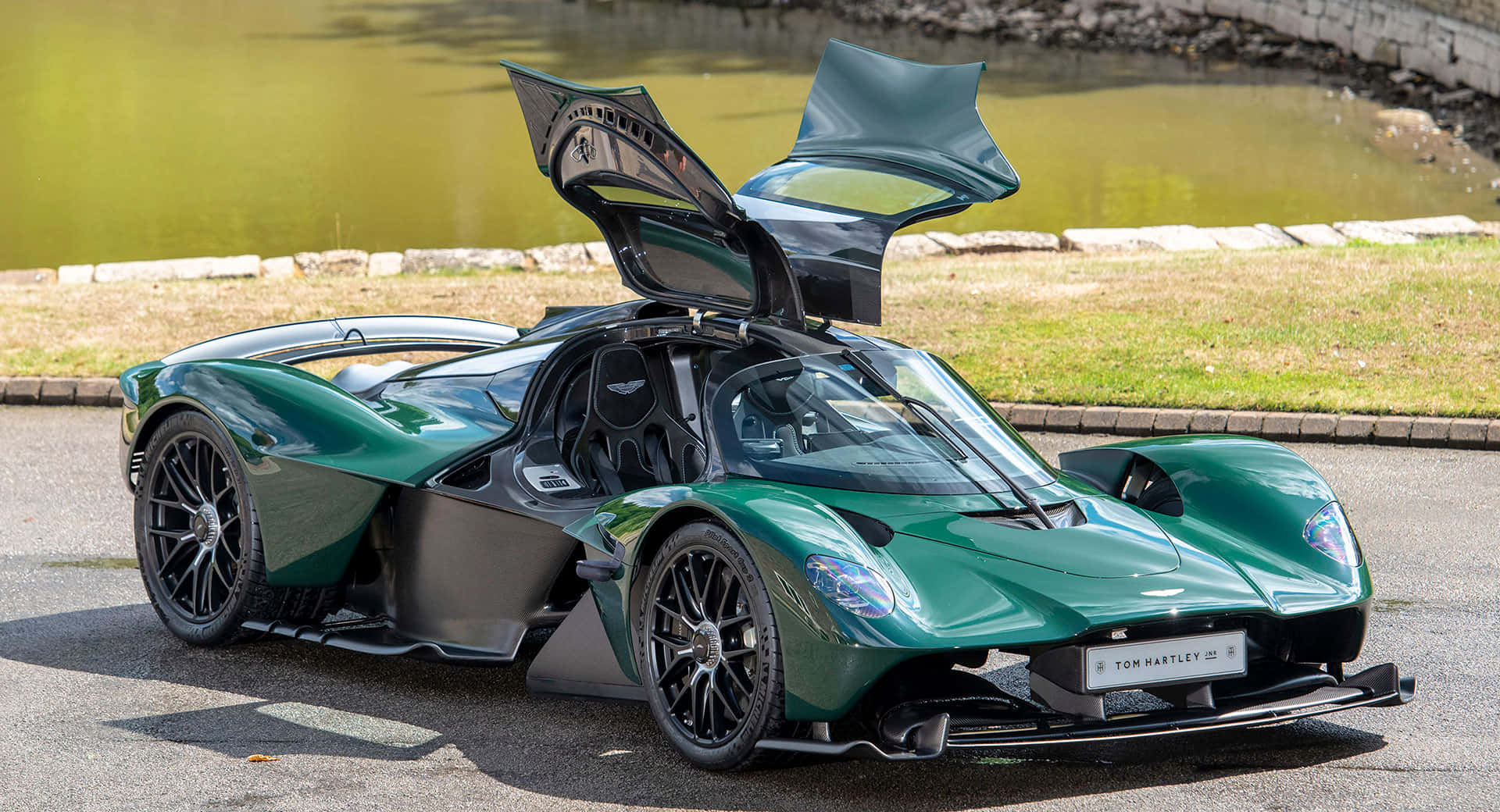 奢華、力量與美學於 Aston Martin 結合 壁紙 — 一架時尚的綠色 Aston Martin Valkyrie 超跑，鷗翼車門打開，停泊在陽光明媚的池塘旁邊的瀝青路上。