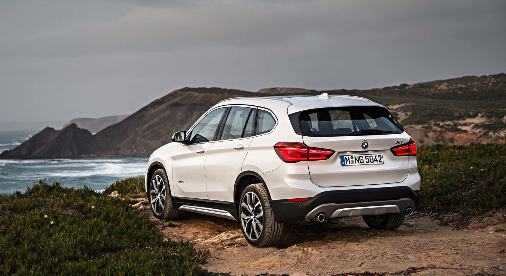 奢華BMW X1駛於風景優美的山道 壁紙 — 一輛白色BMW X1 SUV停泊在崎嶇的海岸懸崖上，俯瞰著下方波濤洶湧的海洋。