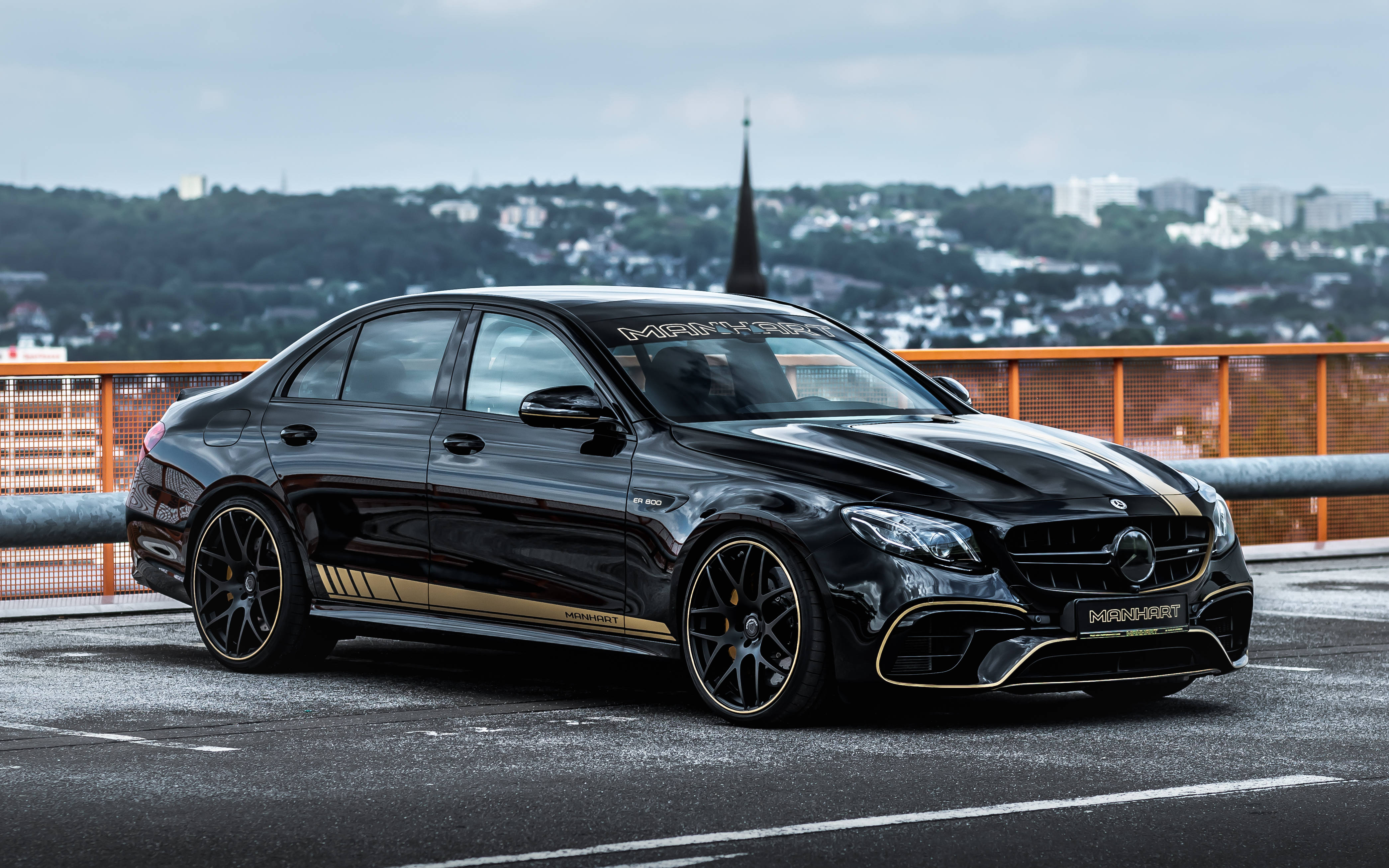 奢華優雅 - 高光黑AMG配橙色圍欄 壁紙 — 一架時尚的黑色 Mercedes-AMG E63 S 帶有金色點綴，停泊在橋上，俯瞰陰天下的城市景觀。