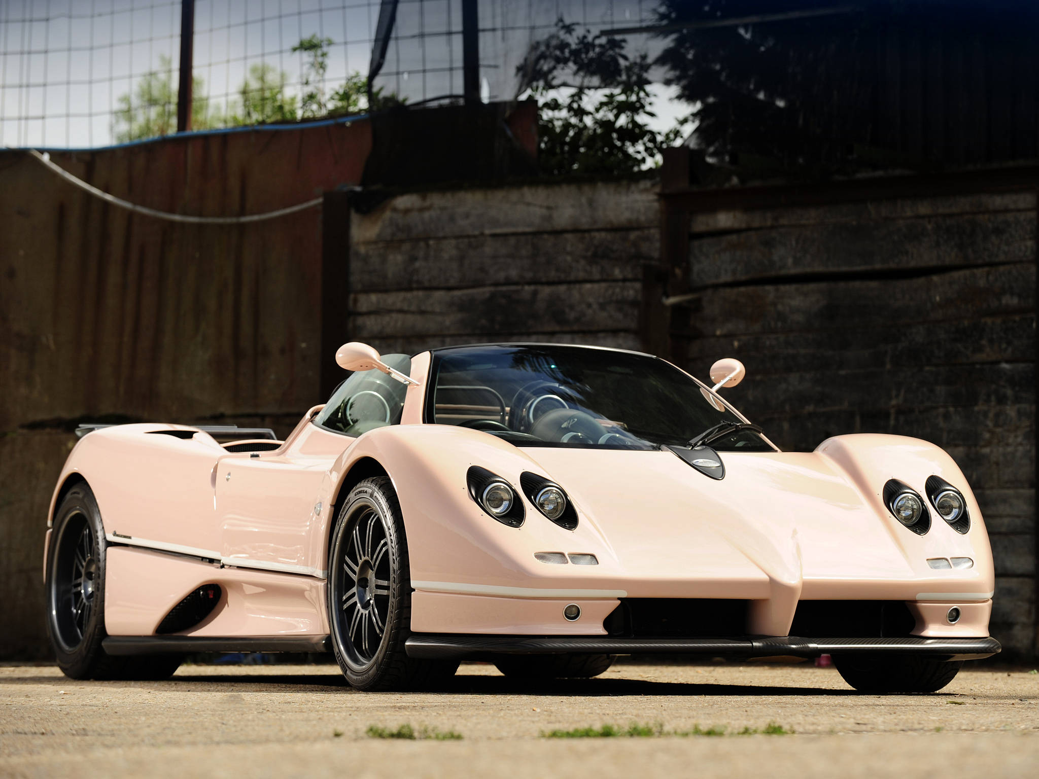 奢華揭幕 — 奶油色 Pagani Zonda C12-S 壁紙 — 一輛時尚的淡粉紅色Pagani Zonda跑車停在混凝土上，背景有木柵欄和綠植。