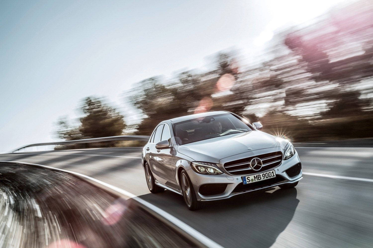 銀色 Mercedes 房車在彎曲道路上疾馳，帶有模糊樹木和鏡頭光暈。