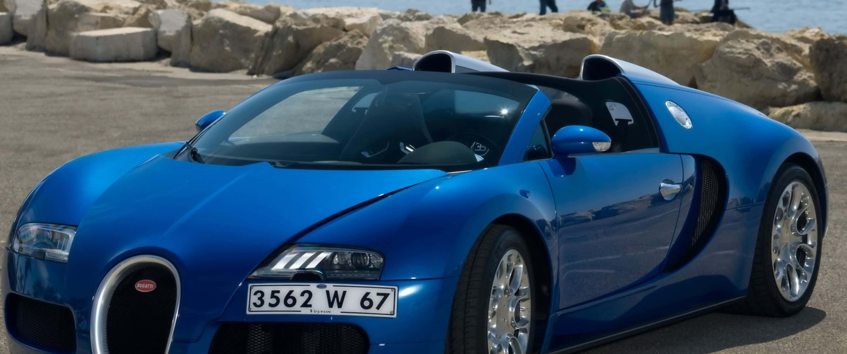 奢華汽車，官方賓利 壁紙 — 一輛充滿活力的藍色Bugatti Veyron超級跑車在明亮的陽光下閃閃發光，停泊在岩石海邊懸崖附近。