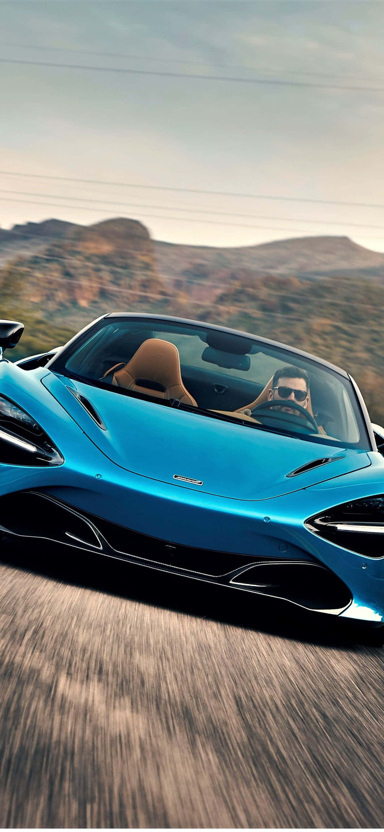 奢華的 McLaren 720S 與 iPhone XS Max 壁紙 — 一輛時尚的藍色跑車在沙漠公路上疾馳，司機戴著太陽眼鏡，背景是崎嶇的山脈。