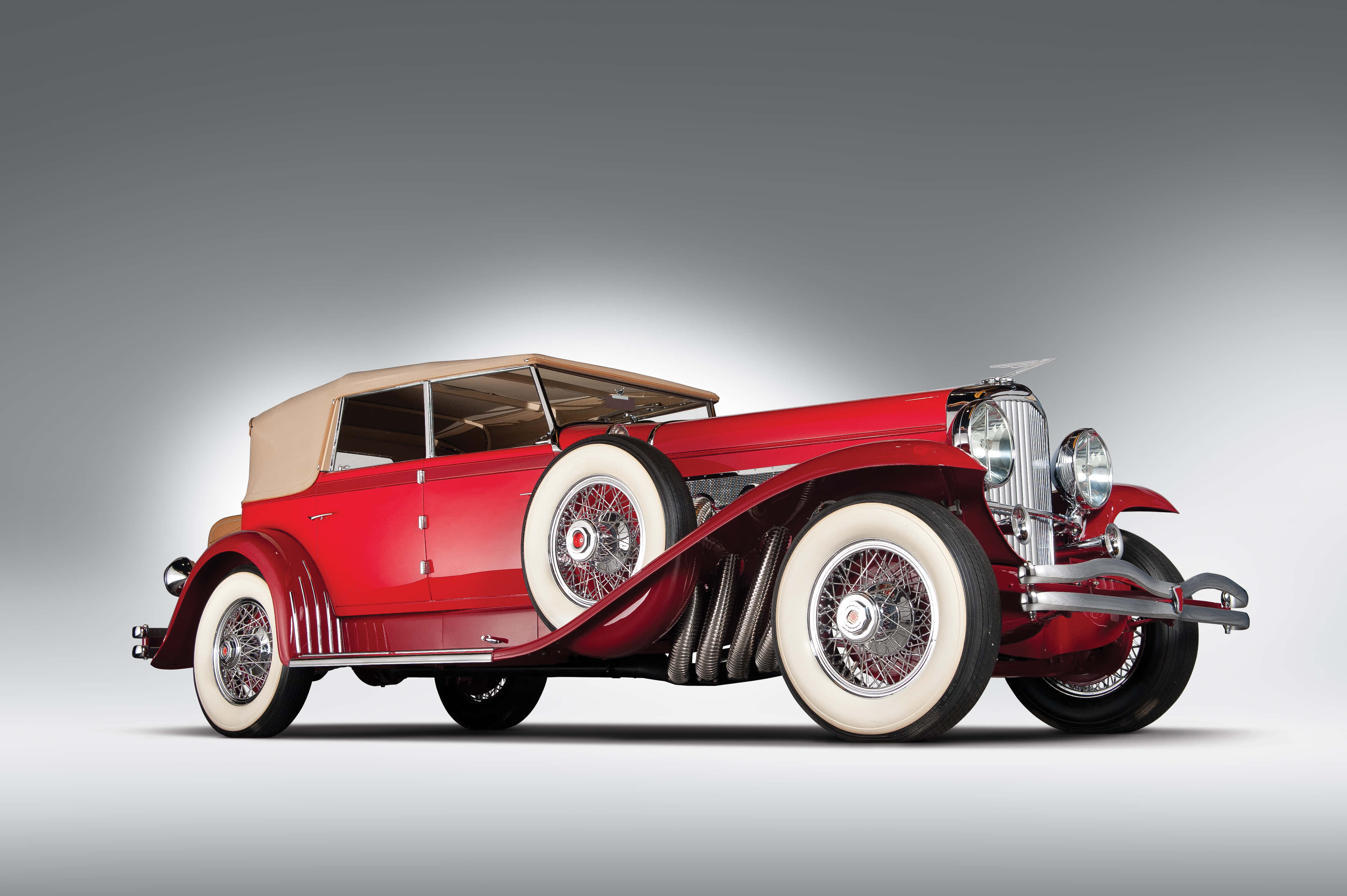 奢華的Duesenberg Model J - 古董豪華車的典範 壁紙 — 一輛光澤的紅色復古敞篷車，配奶油色車頂和白色輪胎，在漸變灰色工作室背景前蓄勢待發。