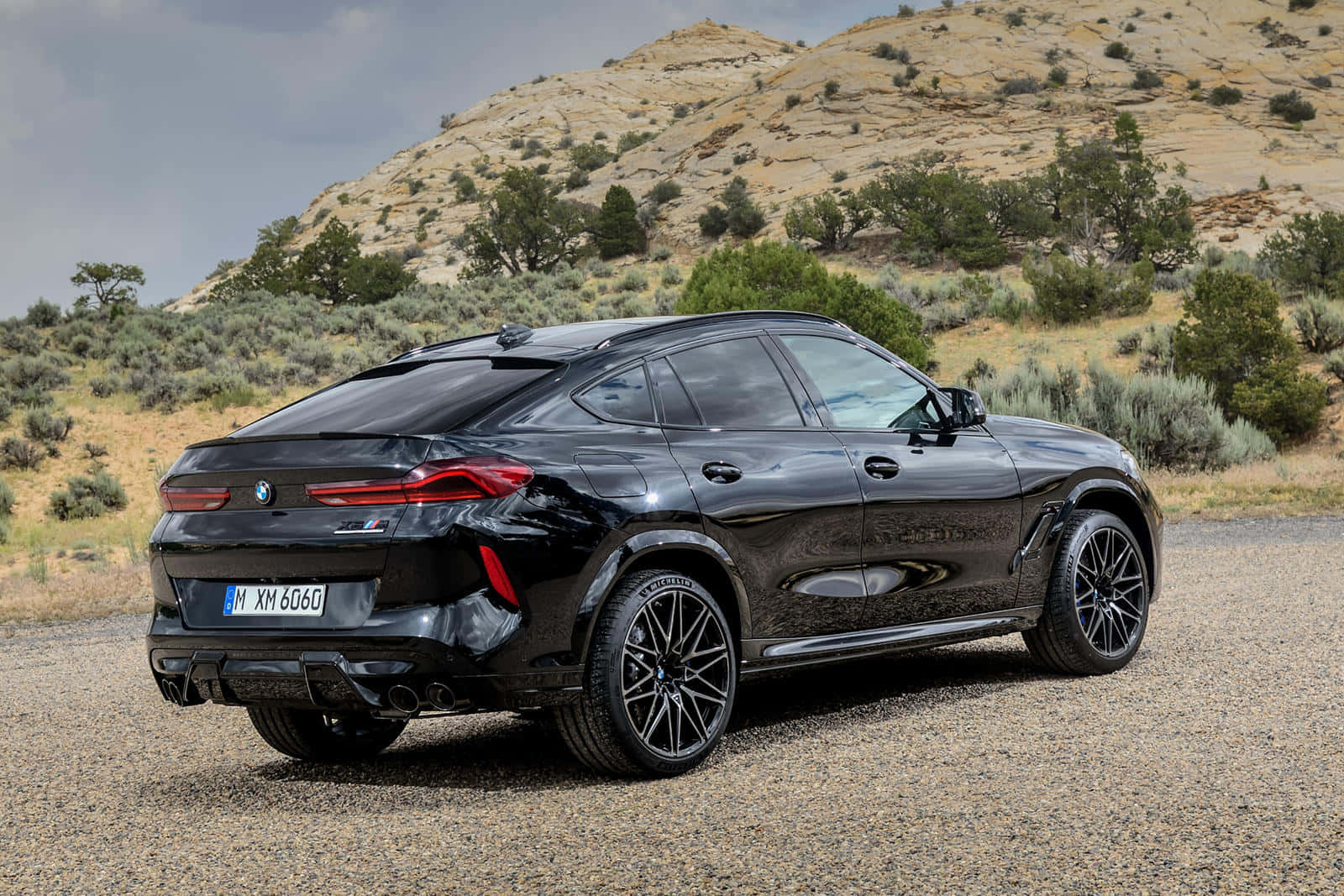 奢華經典黑 BMW X6 M 壁紙 — 一架型格黑色BMW XM SUV停泊在碎石上，背景是乾旱的山丘和稀疏的綠植，天空多雲。