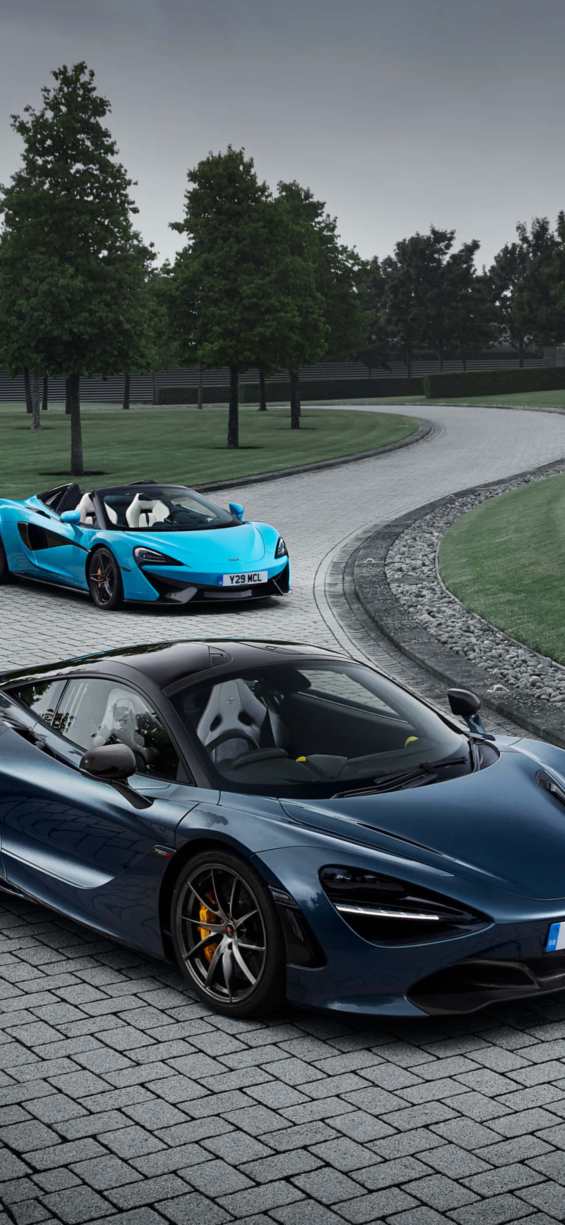 奢華與力量的完美結合：iPhone Xs McLaren 720S 壁紙 — 兩輛時尚的McLaren跑車，一輛藍色開篷車和一輛深藍色轎跑車，停泊在彎曲的石頭車道上，周圍綠樹成蔭，天空多雲。
