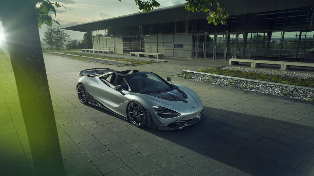 奢華運動型麥拉倫720S 720p 壁紙 — 一架時尚的銀色McLaren Speedtail開篷跑車停泊在現代玻璃建築旁邊的石頭廣場上，陽光穿過樹葉灑落。