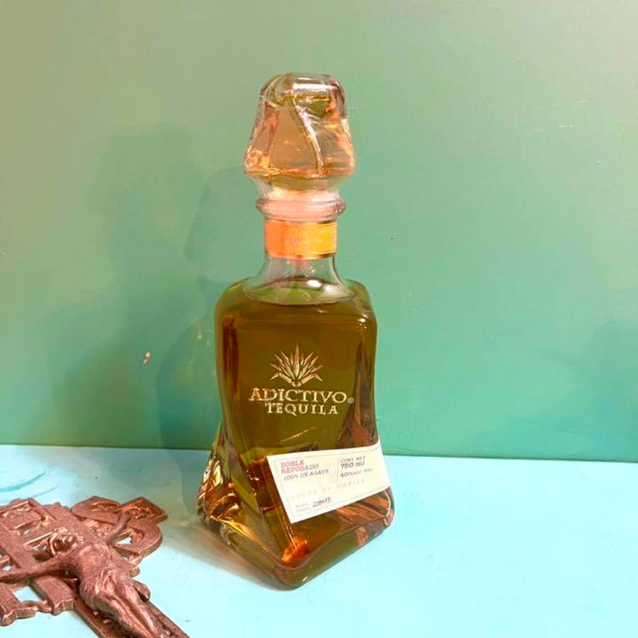 奢華阿迪克蒂沃特級陳年龍舌蘭瓶 壁紙 — 一個有切割面的Adictivo Tequila酒瓶放在青綠色表面上，旁邊有一個青銅十字架，背景是薄荷綠色的牆壁。
