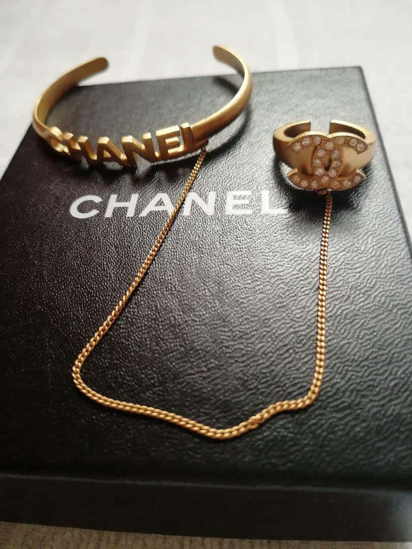 奢華香奈兒標誌 壁紙 — 金色Chanel珠寶首飾，包括標誌手鐲、戒指和鏈條，優雅地放置在黑色紋理Chanel盒子上。