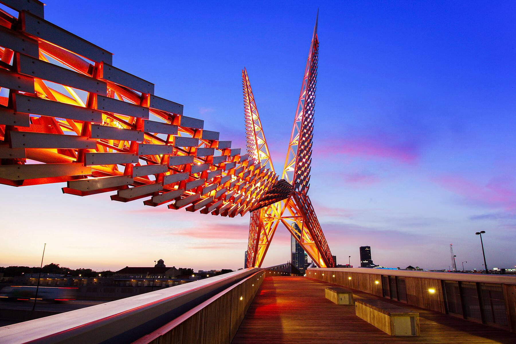 奧克拉荷馬州 Skydance Bridge 夕照景觀 壁紙 — 一座引人注目的現代橋樑，帶有發光的橙色燈光，橫跨城市景觀上空的暮色天空。