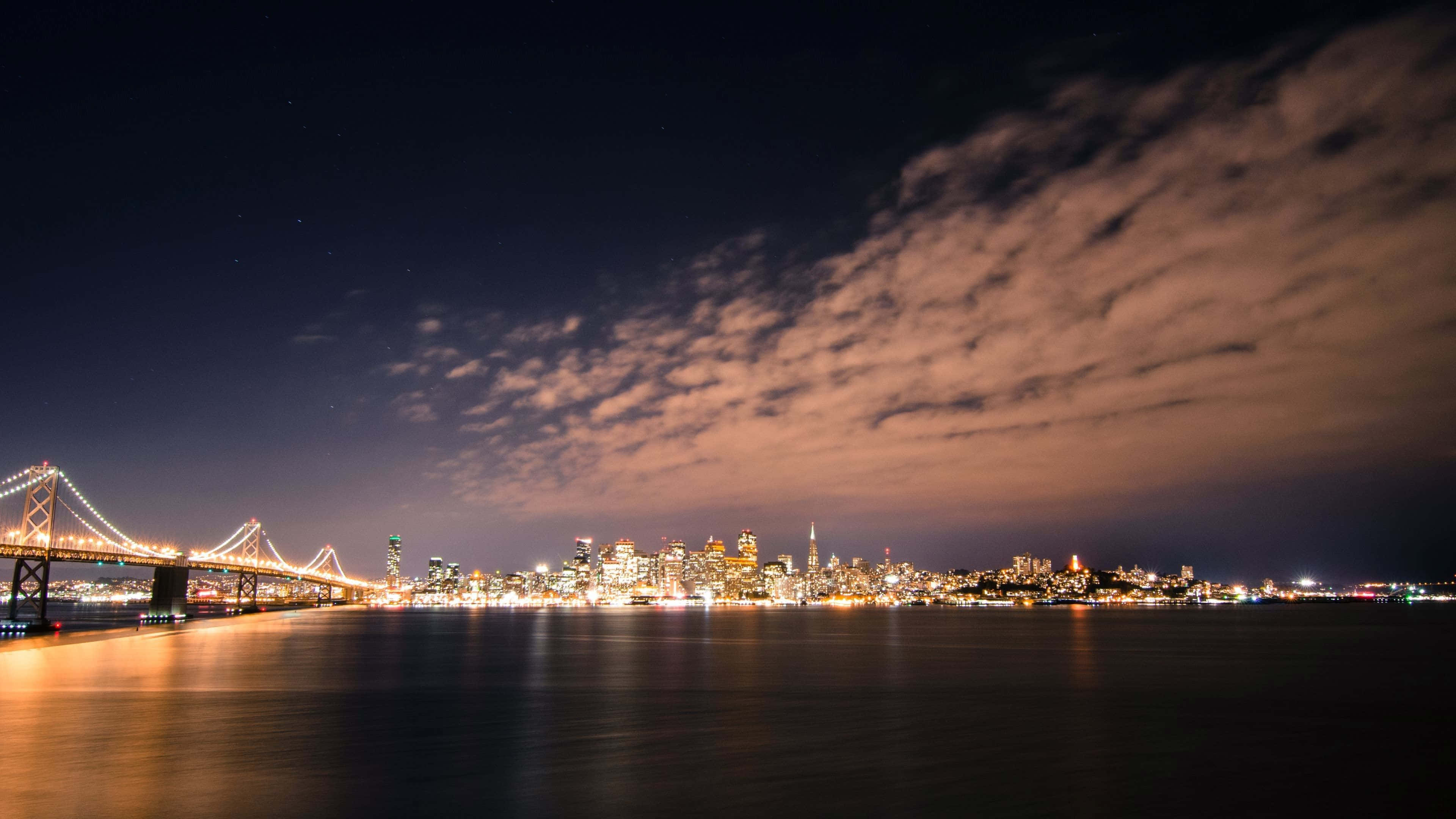 奧克蘭灣橋廣角最佳舊金山背景 壁紙 — 令人驚嘆的城市天際線夜景，一座燈火通明的橋樑橫跨平靜的水面，上方是繁星點點的天空和發光的雲彩。