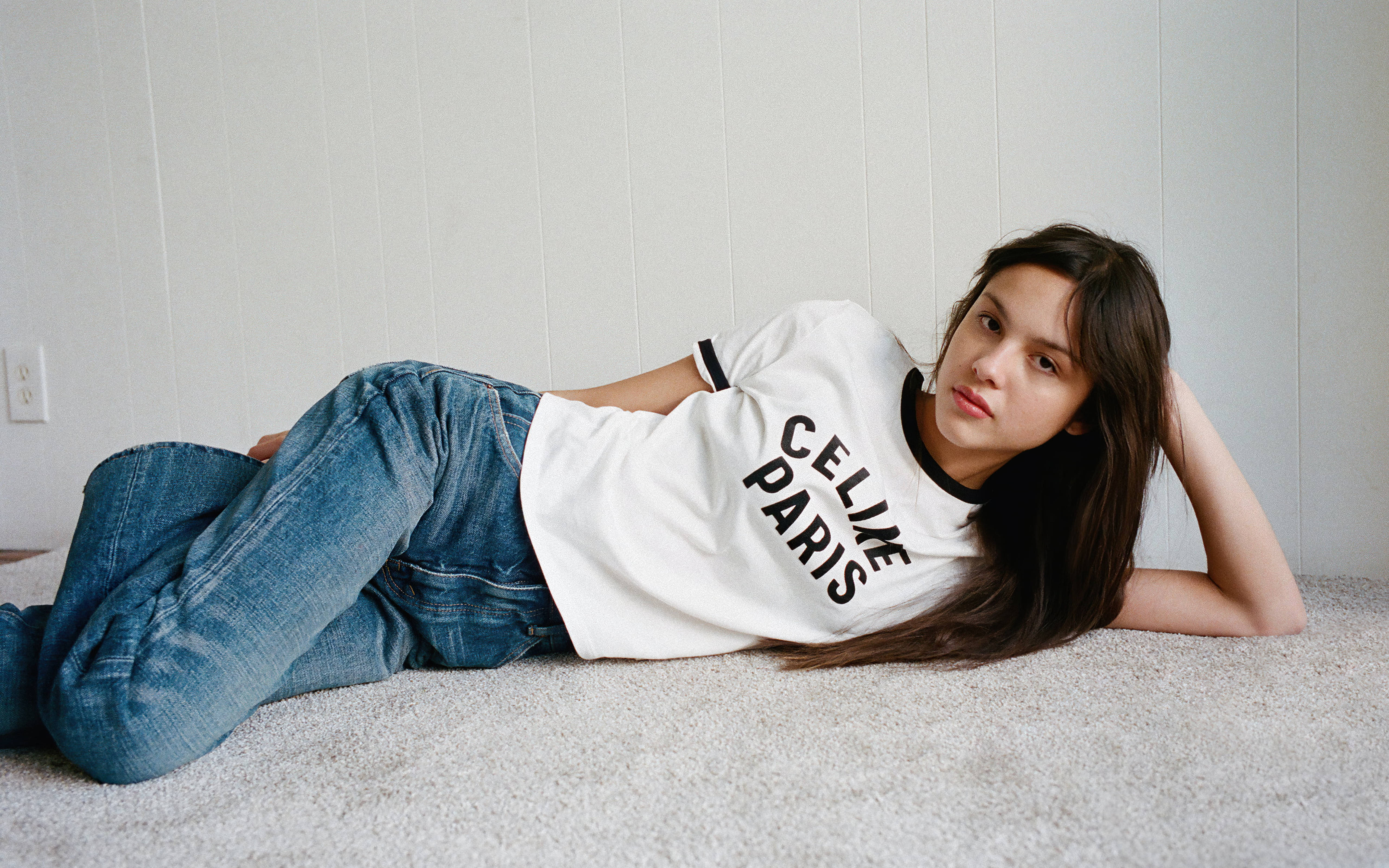 奧利維亞·羅德里戈 W 雜誌 2021 壁紙 — 一位年輕女性穿著白色Celine Paris T恤和藍色牛仔褲，在淺灰色鑲板牆前的米色地毯上悠閒地躺著。