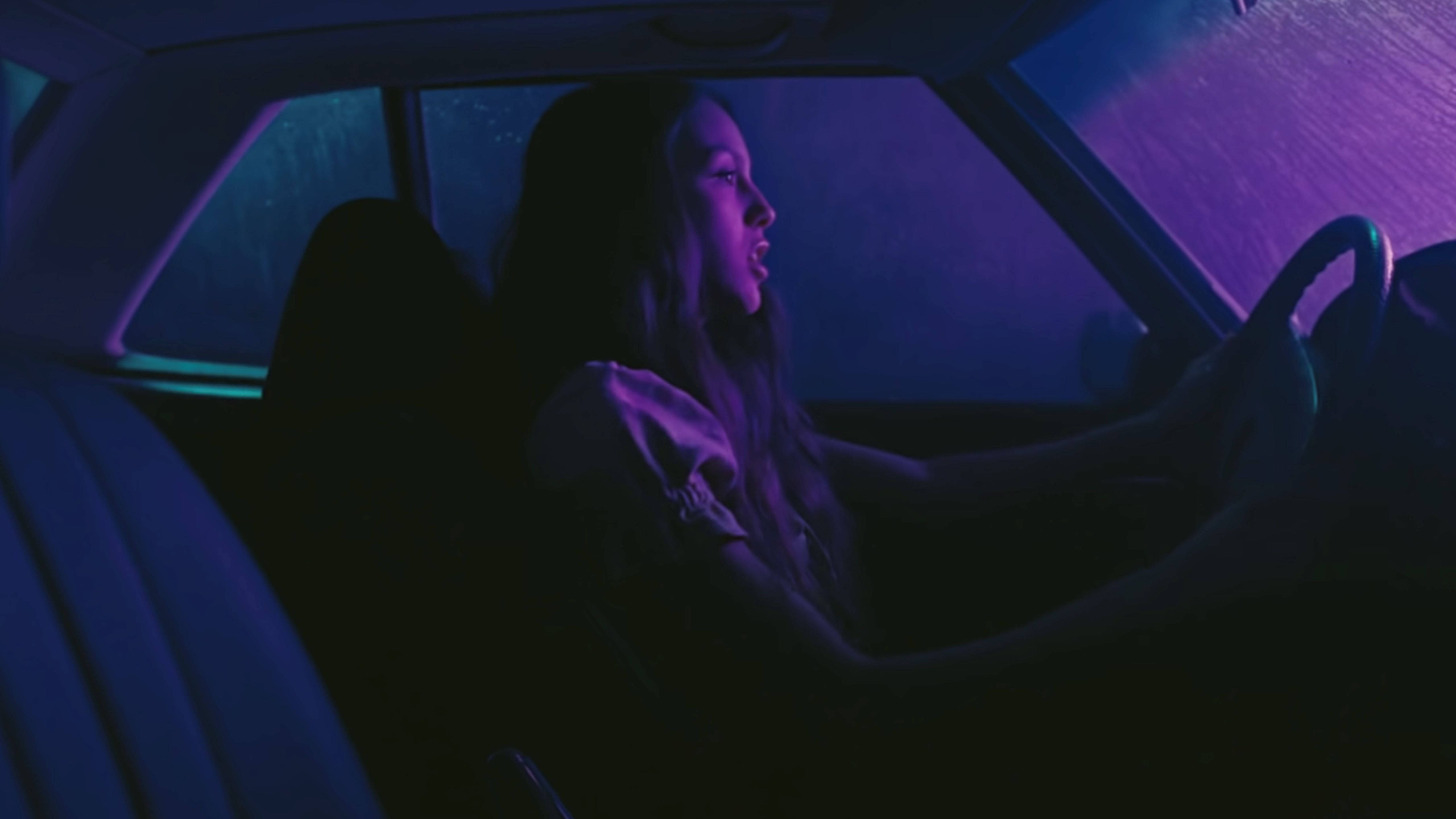 奧利維亞·羅德里戈在《Driver's License》車內場景 壁紙 — 一名女子在夜間駕車穿過雨中的霓虹紫色和藍色街燈，車內視角。