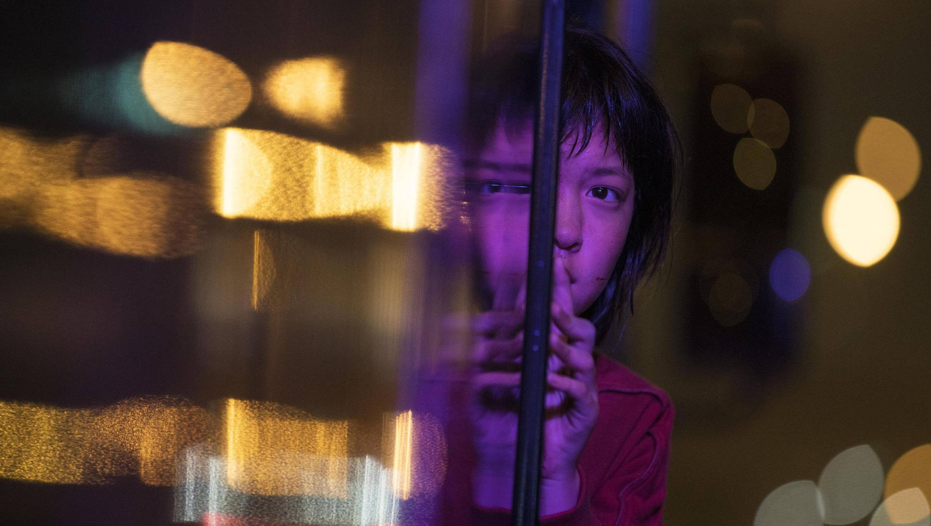 孩子望出被雨水濺濕嘅窗戶，睇到夜晚發光嘅城市燈光。