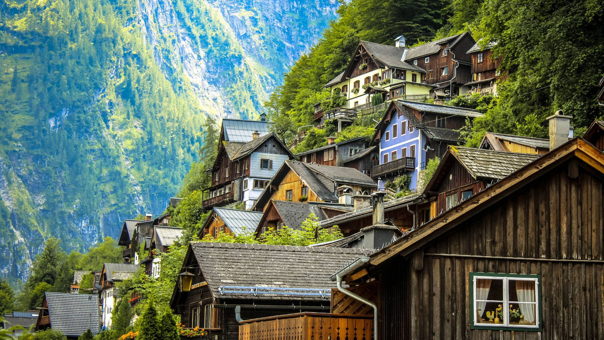 奧地利山坡城市 壁紙 — 色彩繽紛的木屋在明亮的日光下依附在陡峭的森林山坡上。