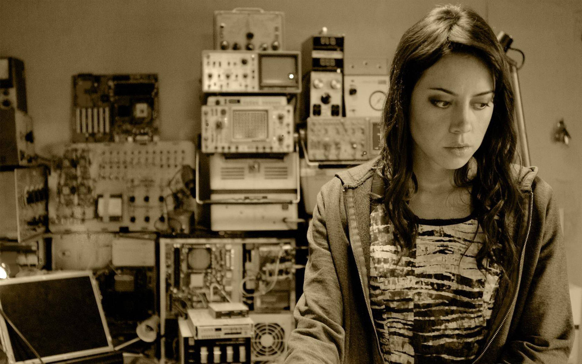 奧布瑞·普拉扎《Safety Not Guaranteed》劇照 壁紙 — 一名穿著連帽衫的女子站在一個雜亂的實驗室中，裡面充滿了復古電子設備，在溫暖的棕褐色燈光下。