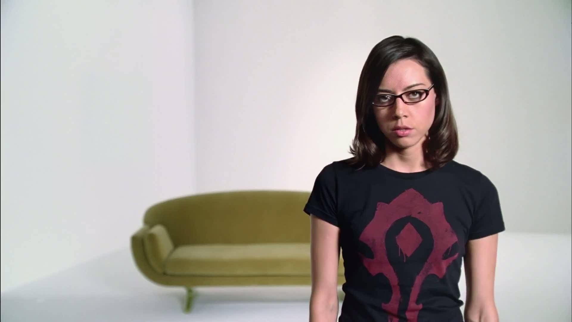 奧布瑞·普拉扎戴眼鏡拍攝 壁紙 — 一位戴眼鏡的女性，在一個簡約的白色房間裡，穿著一件帶有紅色遊戲標誌的黑色T恤，房間裡有一張綠色沙發。