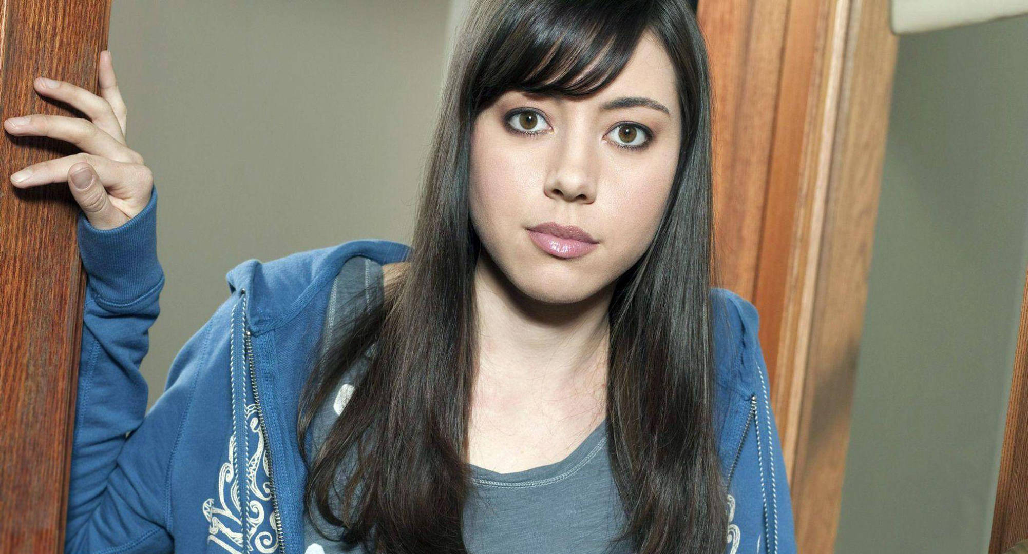 奧布瑞·普拉扎飾演April Ludgate 壁紙 — 一位留著長黑髮和瀏海的年輕女性穿著藍色連帽衫，靠著木門框直視鏡頭。